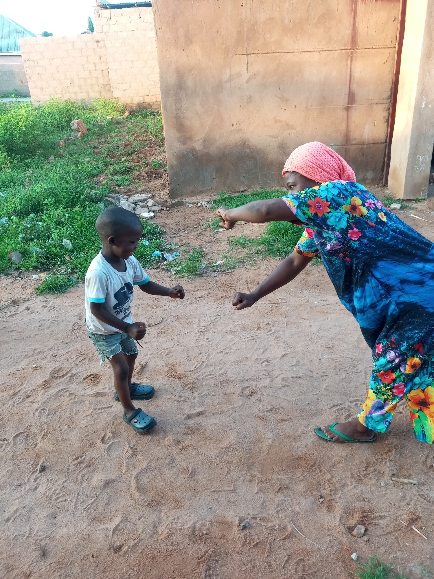 In Tempure, Nigeria, a young boy wearing a white t-shirt, denim shorts, and dark clogs stands on sandy ground. He is looking towards an adult woman, who is wearing a pink headscarf and a vibrant, floral-patterned dress. The woman is facing the boy, with her right arm extended and her fist clenched, appearing to be pointing or gesturing towards him. The background consists of a rough, textured wall and some overgrown vegetation. The scene is outdoors, under natural light, with the ground covered in fine sand and scattered debris.
