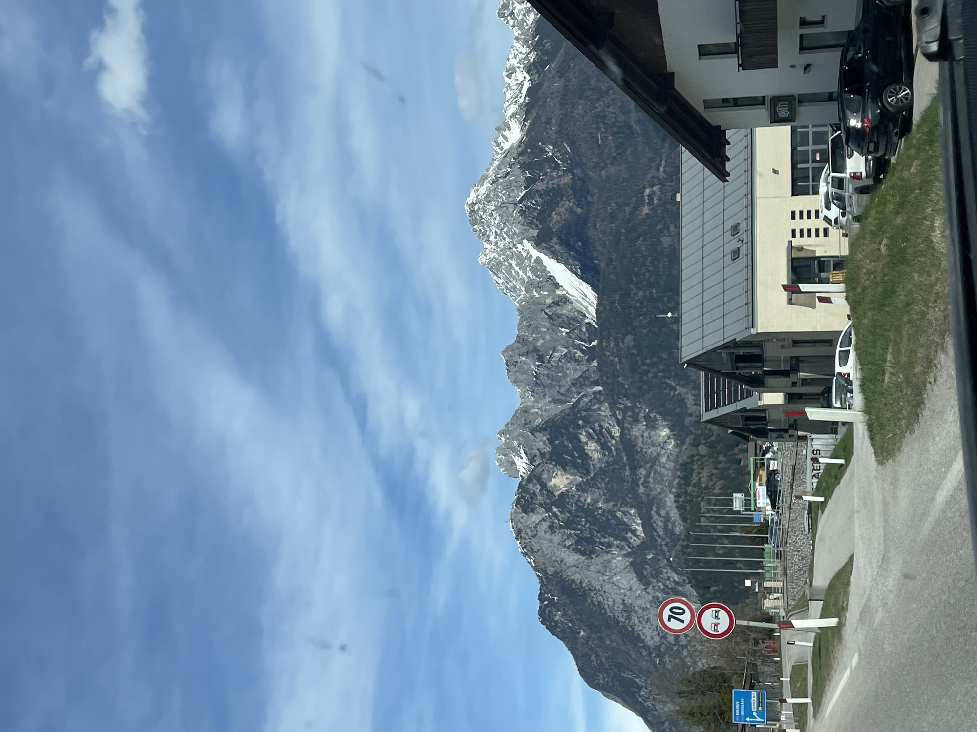 The image shows a view from a vehicle in Lozzo di Cadore, Italy. A mountain range is visible on the left side of the frame, with snow-capped peaks. The sky is blue with scattered clouds. On the right side of the frame, there's a road with a speed limit sign indicating 70 km/h and a sign prohibiting vehicles over 3.5 tons. Buildings and parked cars are present along the road. Grass and other roadside vegetation are also visible. The view suggests the vehicle is traveling along a road near the base of the mountains.