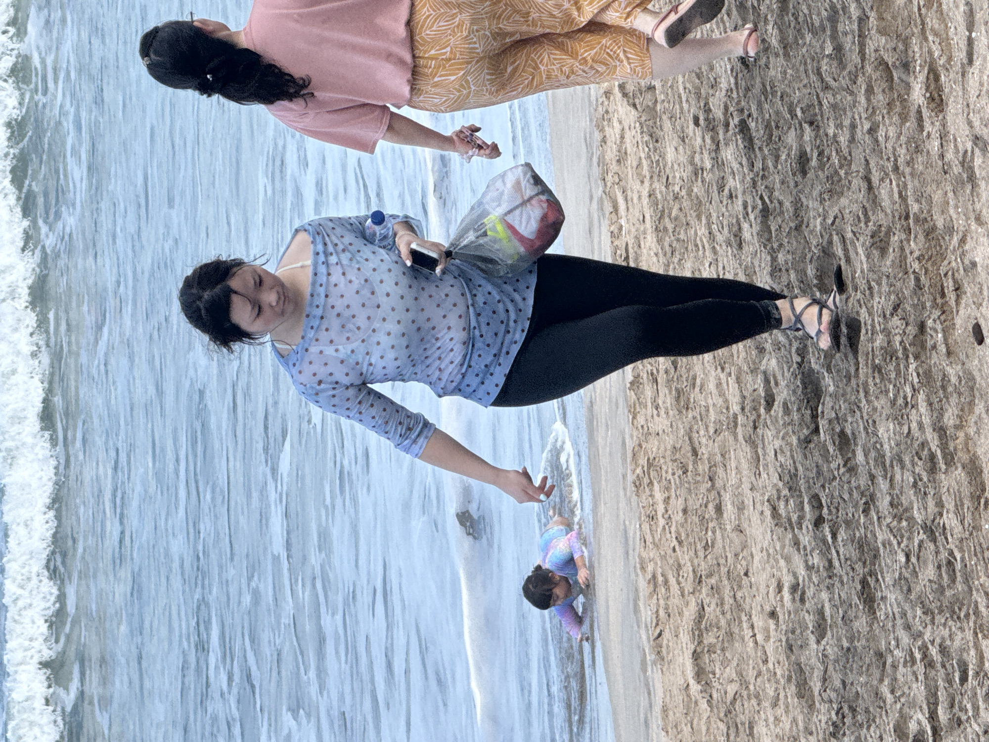 In Kuta, Bali, Indonesia, on a sandy beach, two women walk along the shoreline. The woman on the left wears a sheer blue polka-dot top over a white bra, black leggings, and sandals. She carries a transparent plastic bag filled with items and holds a smartphone in her right hand. The woman on the right, further from the camera, wears a pink t-shirt and patterned yellow pants. She has her hair pulled back and wears glasses. In the shallow water near the shore, a young child crawls, wearing a colorful swimsuit. The ocean is visible in the background with gentle waves.