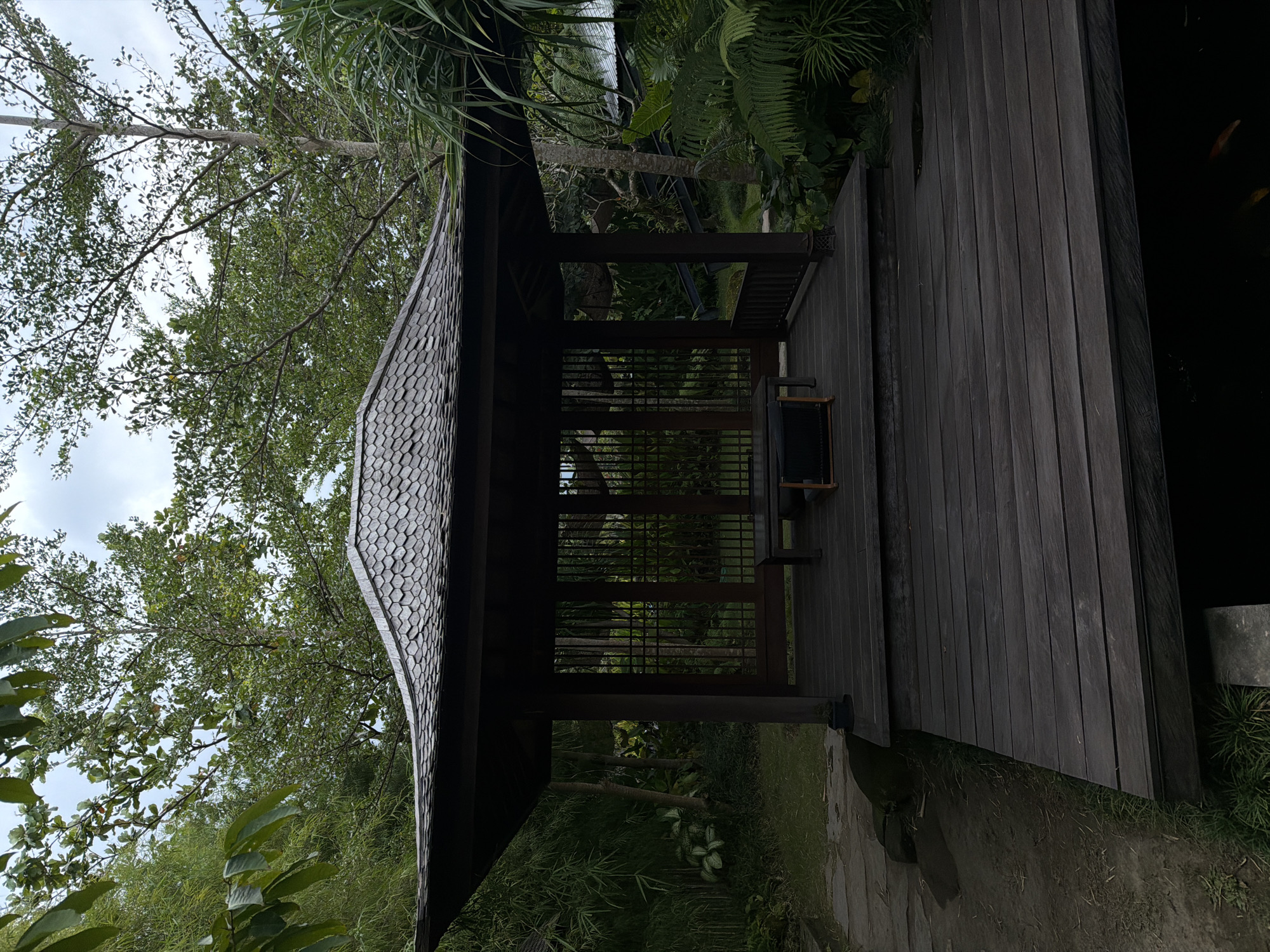 A traditional wooden pavilion stands in a lush tropical garden in Singakerta, Indonesia. The structure features a dark timber frame, a multi-tiered, shingled roof composed of gray overlapping tiles, and an elevated dark wooden plank deck. The pavilion's interior contains a dark wooden table and two low, dark-colored chairs or benches. The sides of the pavilion are partially open, with some sections featuring vertical wooden slat panels.
The deck extends over a dark body of water, likely a pond, where indistinct orange-gold fish are visible near the water's surface. To the left of the pavilion, a dirt path with irregular stone steps or slabs winds through dense greenery, including bamboo stalks. Surrounding the pavilion is abundant tropical foliage, consisting of various trees with green leaves, palm-like plants, and ferns. The sky above is overcast, indicating a cloudy day, and contributes to the soft, somewhat muted lighting of the scene. No individuals are present in the image.