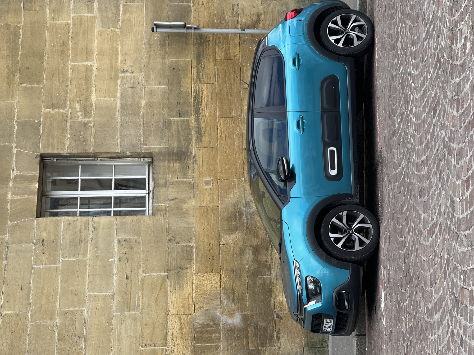 A teal Citroen C3 Aircross is parked parallel to a stone building with a small, square window. The building appears to be an older structure with rectangular stone blocks. A silver light fixture extends from the building above the car. The ground is paved with a herringbone pattern of reddish-brown cobblestones. The license plate of the car is visible. The image suggests a street scene in a European city. The location is Metz, France.