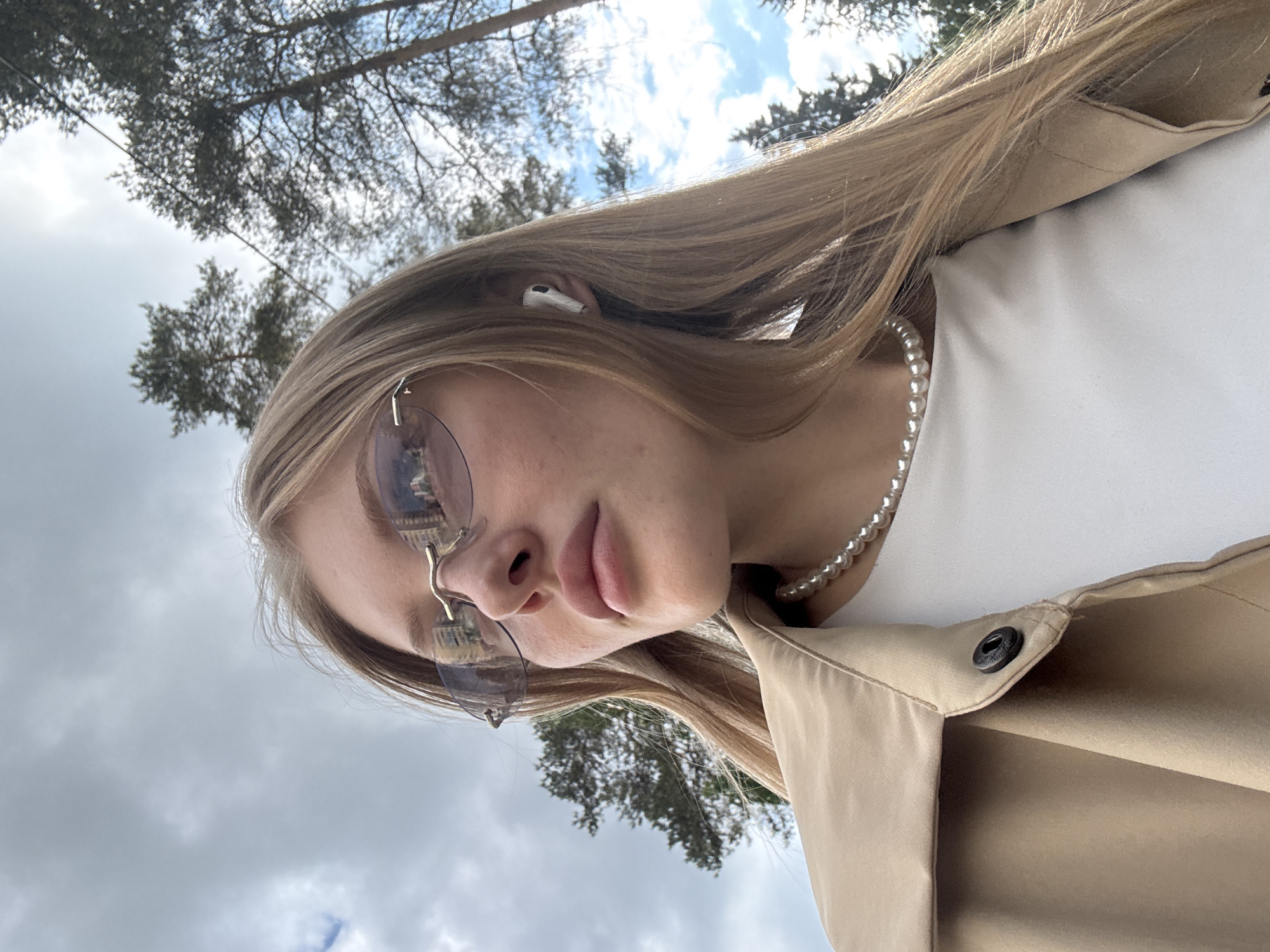 A close-up, portrait-style photograph captures a young woman outdoors, rotated 90 degrees clockwise from its natural orientation. If viewed upright, the woman's head is tilted, and she is looking towards the upper right of the frame. She has long, straight blonde or light brown hair that cascades around her face and shoulders.
She is wearing sunglasses with lightly tinted lenses and a minimalist gold frame; reflections in the lenses show what appear to be architectural structures, possibly buildings or a dome. A white AirPod is visible in her left ear, partially covered by her hair. Around her neck is a pearl necklace. She wears a beige trench coat or jacket with a visible black snap button detail on the lapel, layered over a simple white top. Her lips are slightly parted, and her expression appears neutral or contemplative.
The background, seen from a low angle, consists of tall, slender trees, likely conifers such as pines, their branches silhouetted against a largely cloudy sky. Some patches of blue sky are visible through the canopy. The weather appears overcast or partly cloudy, indicating daytime. The setting suggests a park, forest, or natural area in or near Perm, Russia. No visible text is present in the image.