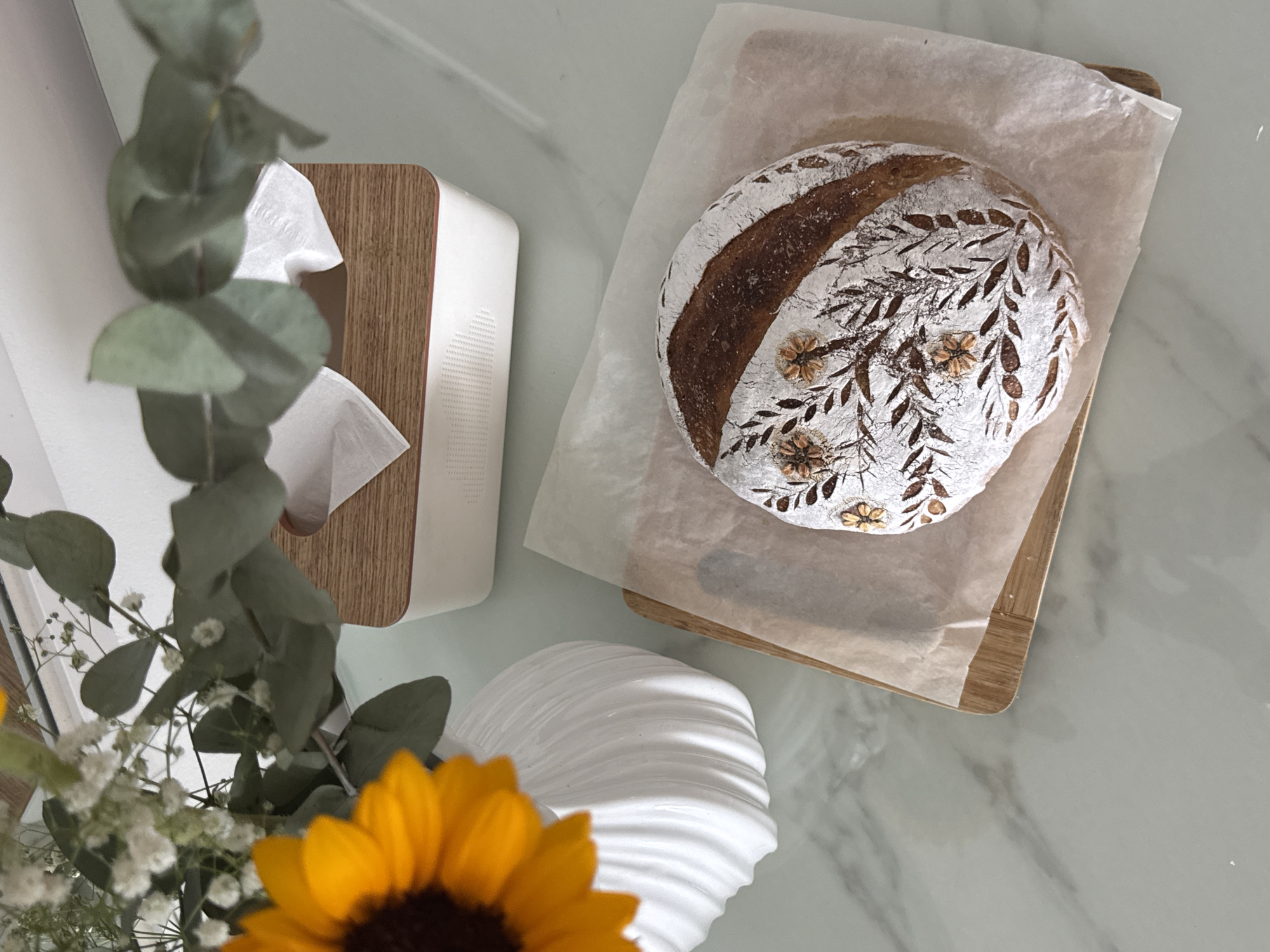 This is a top-down, eye-level shot of a decorative sourdough loaf of bread positioned on a wooden cutting board lined with parchment paper. The bread is centrally located, featuring intricate scoring that resembles flowers and leaves, dusted with flour. To the left of the bread, a white, textured vase holds a sunflower and a bouquet of baby's breath and eucalyptus leaves. Behind the vase and to the right of the bread is a white and wood-toned tissue box. The scene is set on a light gray, marble-patterned surface. The image does not depict the city of Bac Nha Trang Ward, Vietnam.