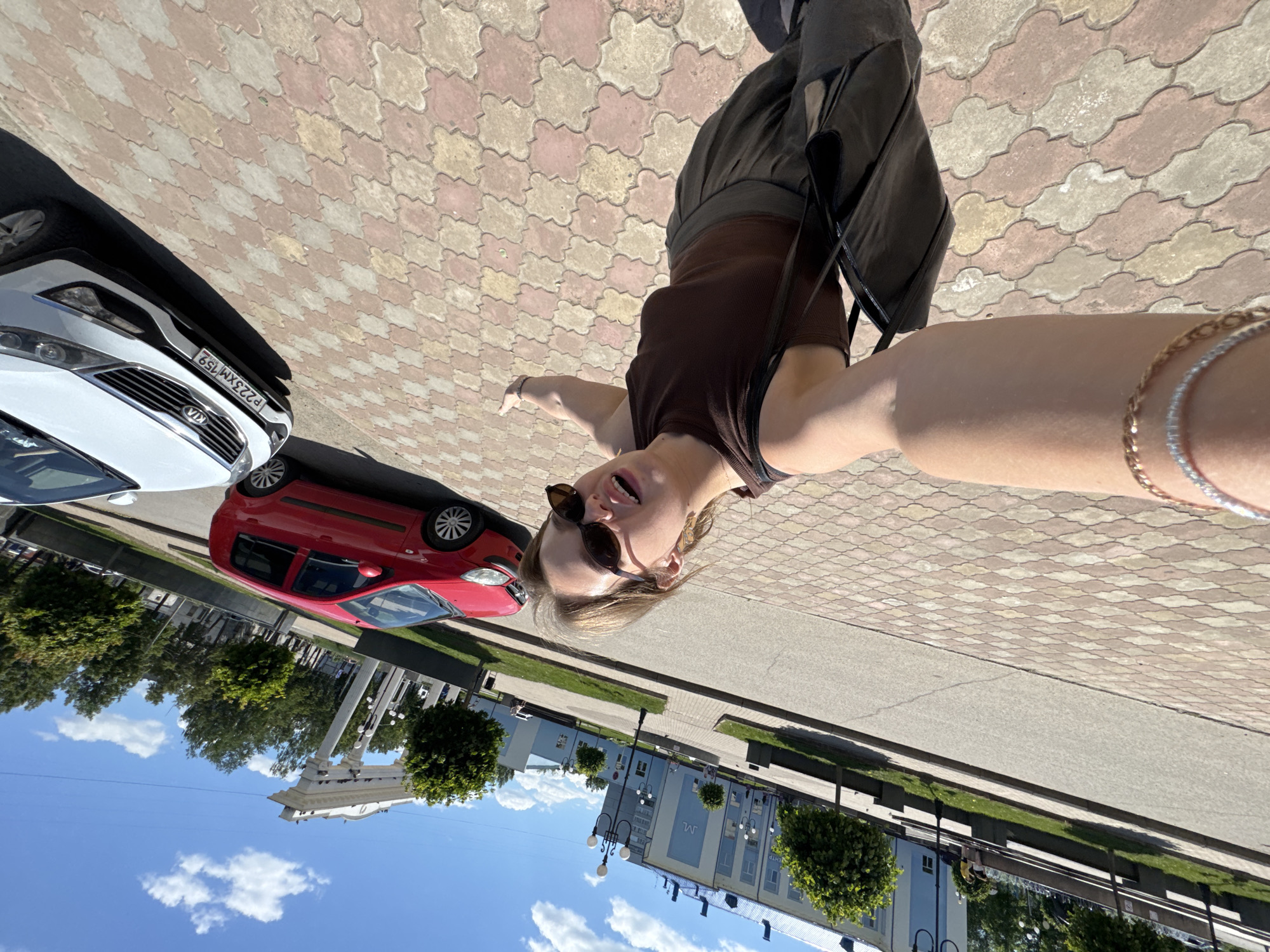 The image shows a young woman taking a selfie on a sunny day in Chaikovsky, Russia. She is wearing sunglasses, a brown tank top, and grey pants, and is carrying a black bag over her shoulder. Her arm is adorned with bracelets. She is standing on a sidewalk paved with interlocking red and beige tiles. In the background, there are two cars parked on the street: a red hatchback and a white SUV with a license plate visible. Behind the cars, a light blue building with white trim and a dark blue roof can be seen, along with trees and a white archway structure. The sky is blue with scattered white clouds. The overall scene appears to be a public space in a city, likely a town square or a street near a civic building.