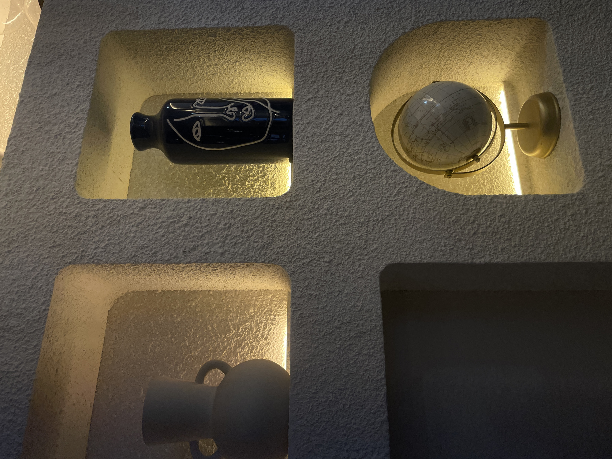 A close-up shot depicts a textured, light-colored wall in Puebla, Mexico, featuring four recessed compartments. The top left compartment contains a horizontally placed, dark-colored bottle with a white, stylized face design. Adjacent to it, in the top right compartment, a world globe is mounted on a brass-colored fixture. The lower left compartment houses a ceramic vase with two handles, while the lower right compartment is empty and dark. Each compartment is illuminated from within, highlighting the objects and the wall's texture.