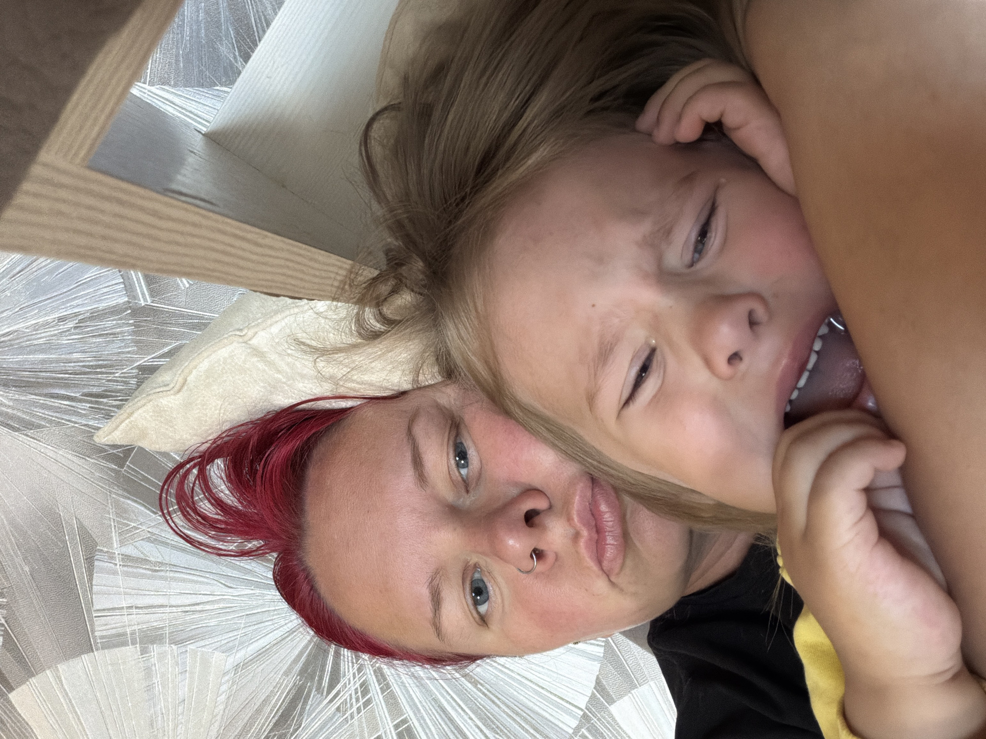 In a close-up, overhead shot taken from a bedroom in Chaikovsky, Russia, a woman with vibrant red hair and a young child are partially visible. The woman is in the foreground, with her face turned towards the camera, and her lips pursed. She is wearing a black t-shirt. To her right, the child's face is angled away from the camera, with their eyes closed and mouth open. The child's hand is visible near their mouth. The background consists of a textured wallpaper with a geometric pattern and a portion of a wooden structure. The scene suggests an intimate moment between the two subjects.