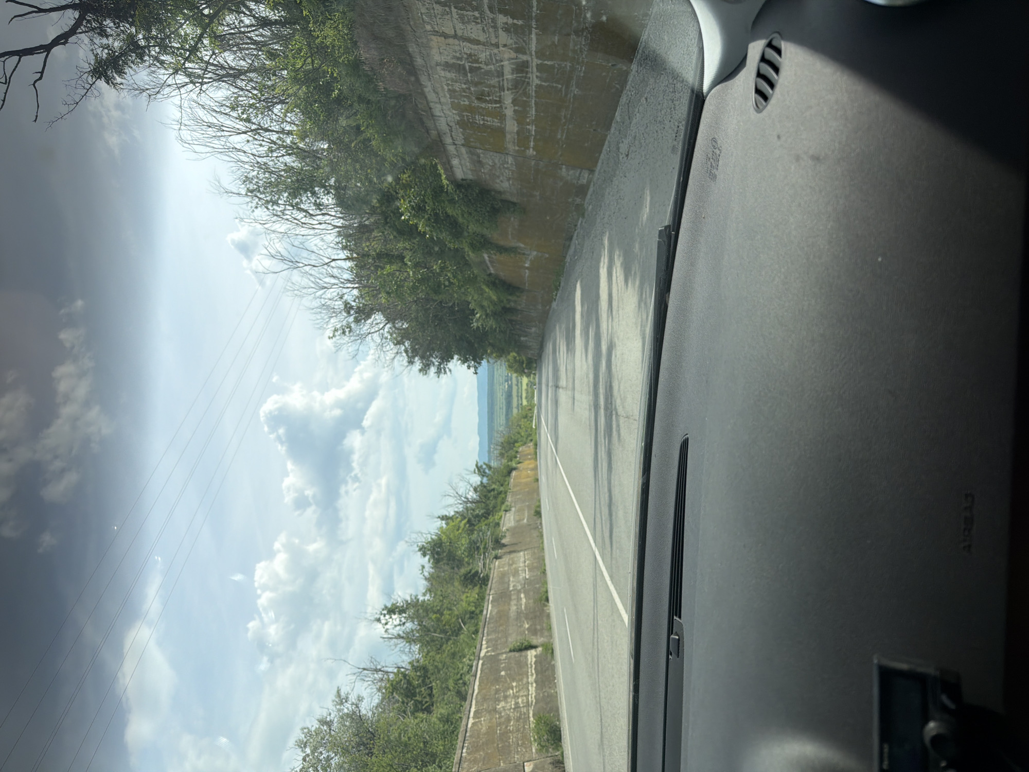 The image captures a view from inside a vehicle, looking forward and slightly to the right through the windshield. The scene depicts a road trip through a varied landscape, likely in the region of Shumen, Bulgaria.
In the foreground on the right, the dark gray dashboard of the car occupies a significant portion of the frame. An air vent with horizontal slats is visible in the upper right. The embossed text "AIRBAG" is discernible on the dashboard surface, oriented sideways. Reflections, including a bright glare from the sun, are visible on the windshield, along with faint streaks or dust. A small electronic device, possibly a dashcam or navigation unit, is partially visible at the bottom right corner of the dashboard.
The paved road extends into the distance, marked by a solid white line on the right edge and a broken white line dividing the lanes or indicating a shoulder. To the left of the road, a tall, weathered concrete retaining wall runs parallel to the vehicle's direction, partly overgrown with lush green foliage and some dry, skeletal trees and bushes. The wall shows signs of wear and moisture, with dark stains and patches of moss.
Beyond the retaining wall and the immediate roadside vegetation, a broad panoramic view of distant fields and rolling hills stretches out. The sky above is dynamic, featuring a mix of bright, sunlit areas with fluffy white clouds and darker, heavier clouds towards the upper left, suggesting a partly cloudy or overcast day. Several thin power lines or utility cables are visible against the sky in the upper left quadrant.
The image conveys a sense of daytime travel, likely late morning or early afternoon, with the shifting light and shadows created by the clouds. No people are visible in the image.