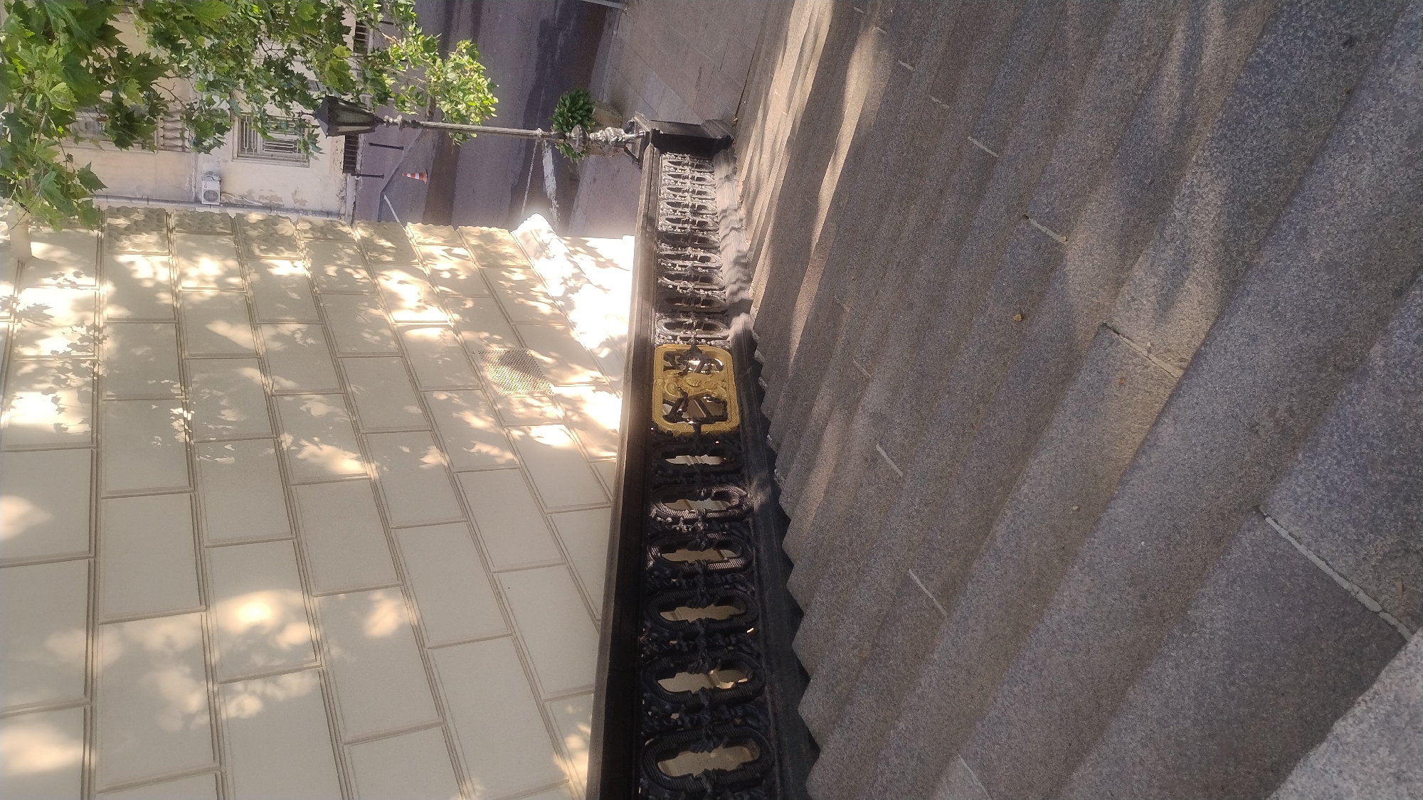 This is a high-angle shot looking down at a textured, light-colored tiled wall next to a set of stone steps. The tiled wall is partially covered in dappled sunlight and shadows, suggesting it is daytime with trees casting shadows. To the left of the tiles, a building with windows and an air conditioning unit is visible, with green tree branches hanging over it. Next to the building, a street can be seen with a lamppost and a traffic cone.
The stone steps are dark gray and appear to be a public walkway or part of a building's exterior. A decorative black metal railing with ornate detailing separates the tiled wall from the steps. A prominent gold-colored decorative element is integrated into the railing, drawing the eye. The overall impression is one of an urban outdoor setting, likely during daylight hours with pleasant weather allowing for natural light to illuminate the scene. There is no visible text.