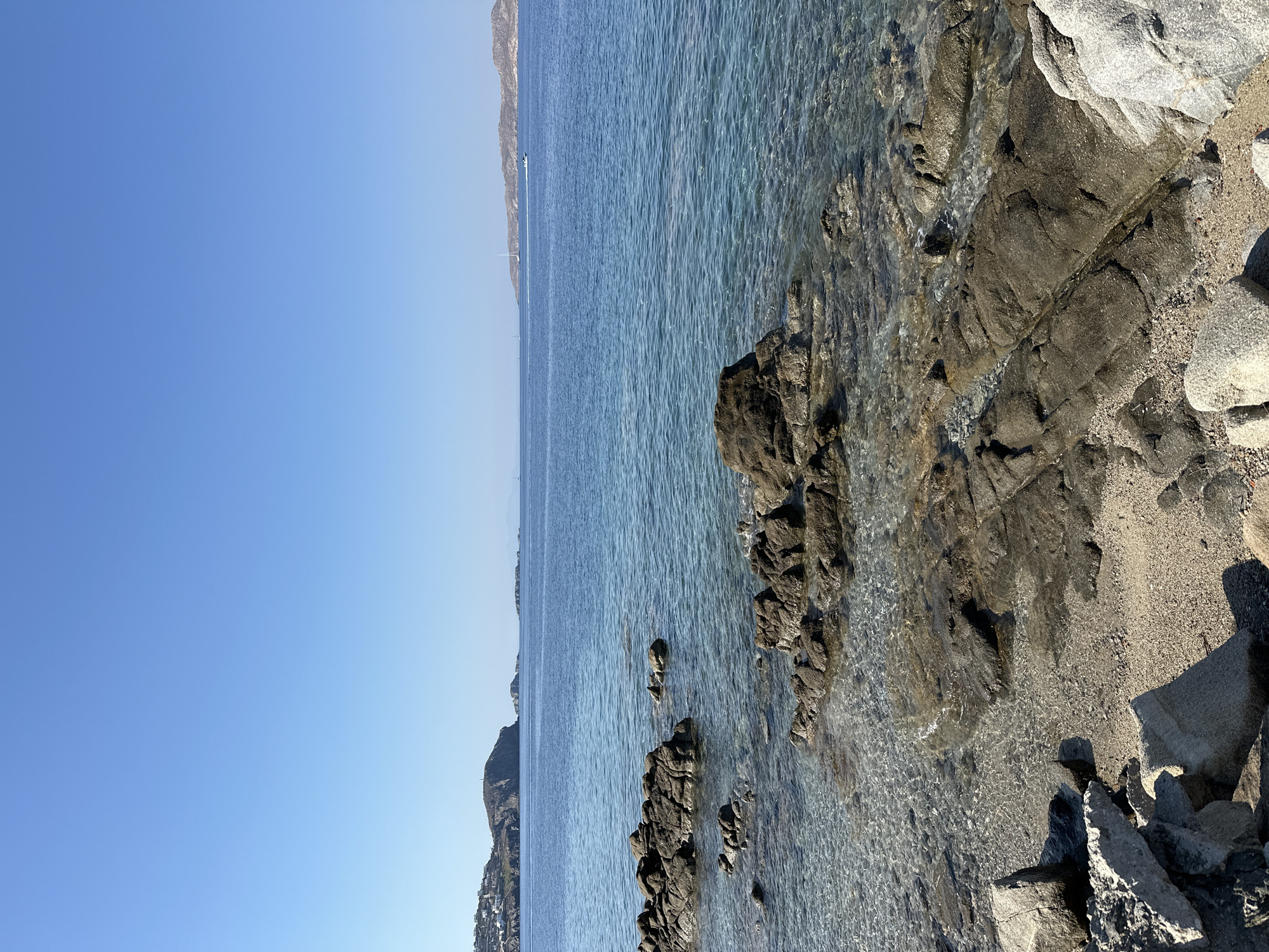 The image depicts a coastal scene in Agios Ioannis Diakoftis, Greece, under a clear blue sky. In the foreground, the rocky shoreline transitions into the transparent, shallow waters of the Aegean Sea. Scattered rocks of various sizes are visible both on the sandy shore and partially submerged in the water. The sea itself is a calm, deep blue, with sunlight creating shimmering patterns on its surface. In the mid-ground, the coastline of a hilly island is visible, dotted with white buildings. Further in the distance, other islands are faintly visible on the horizon, with a few sailboats and a small boat with a wake cutting through the water. The overall impression is one of a serene and picturesque Mediterranean seascape.