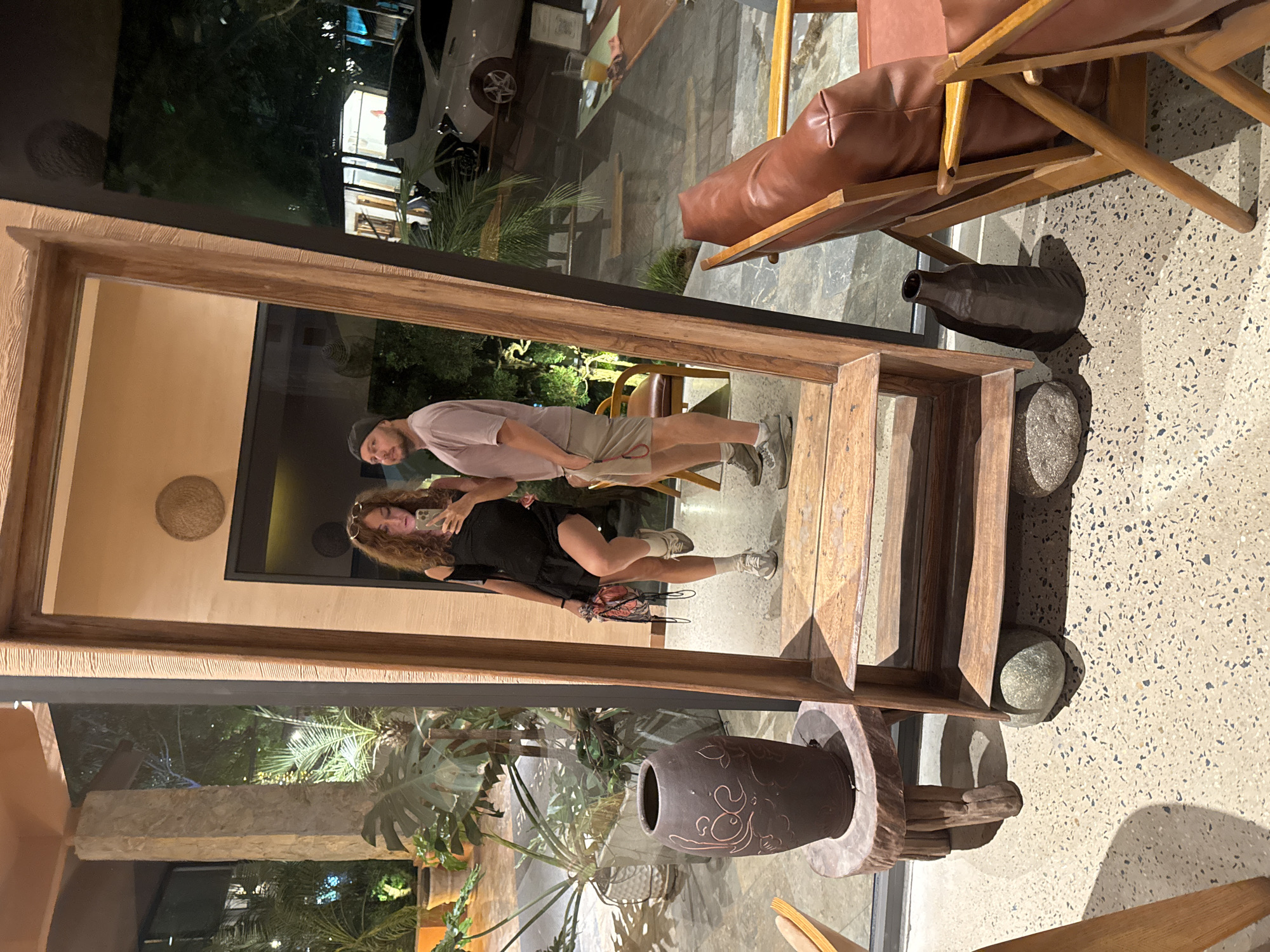 The image shows a young couple taking a mirror selfie in what appears to be an outdoor seating area of a restaurant or cafe, possibly located in Nam Hoa Lư Ward, Vietnam. The woman on the left is wearing a short black dress and holding a red bag. The man on the right is wearing a pink t-shirt and beige shorts, with a dark grey baseball cap. They are standing in front of a large, rustic wooden mirror. In the background, there are large potted plants, stone pillars, and outdoor furniture, including brown leather chairs. A white car is visible through a glass door in the background, along with other tables and chairs. The lighting suggests it is evening or night. The flooring is a terrazzo-like material. A dark brown decorative vase is placed on a wooden stool to the left of the mirror, and another dark bottle-shaped vase is on the floor to the right. Several large grey stones are also visible on the floor, some supporting the mirror stand.