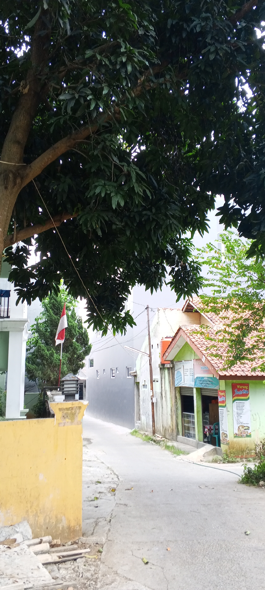 The image captures a street scene in Depok, Indonesia, under a cloudy sky. A large, mature tree with dense green foliage dominates the upper left portion of the frame, its branches casting shadows. In the foreground, a weathered yellow wall partially obstructs the view, with some construction debris near its base. A paved road curves to the right, bordered by buildings.
To the left of the road, a conical green tree stands tall, adorned with a small red and white Indonesian flag atop a pole. Adjacent to this is a white building with a dark balcony railing. Further down the street, a grey building features a repetitive pattern of small, rectangular windows.
On the right side of the road, a small, brightly colored building with a red tiled roof appears to be a shop or warung. It has a green facade with a brown awning over its entrance, displaying various signage, including one that reads "Warung Indomie." A display window reveals shelves stocked with goods, and a green chair is visible inside. The building's exterior walls show signs of age and weathering.
The street itself is made of cracked asphalt, with scattered leaves and small debris. The overall impression is a typical urban residential or commercial street in Indonesia, characterized by a mix of residential structures, small businesses, and abundant greenery.