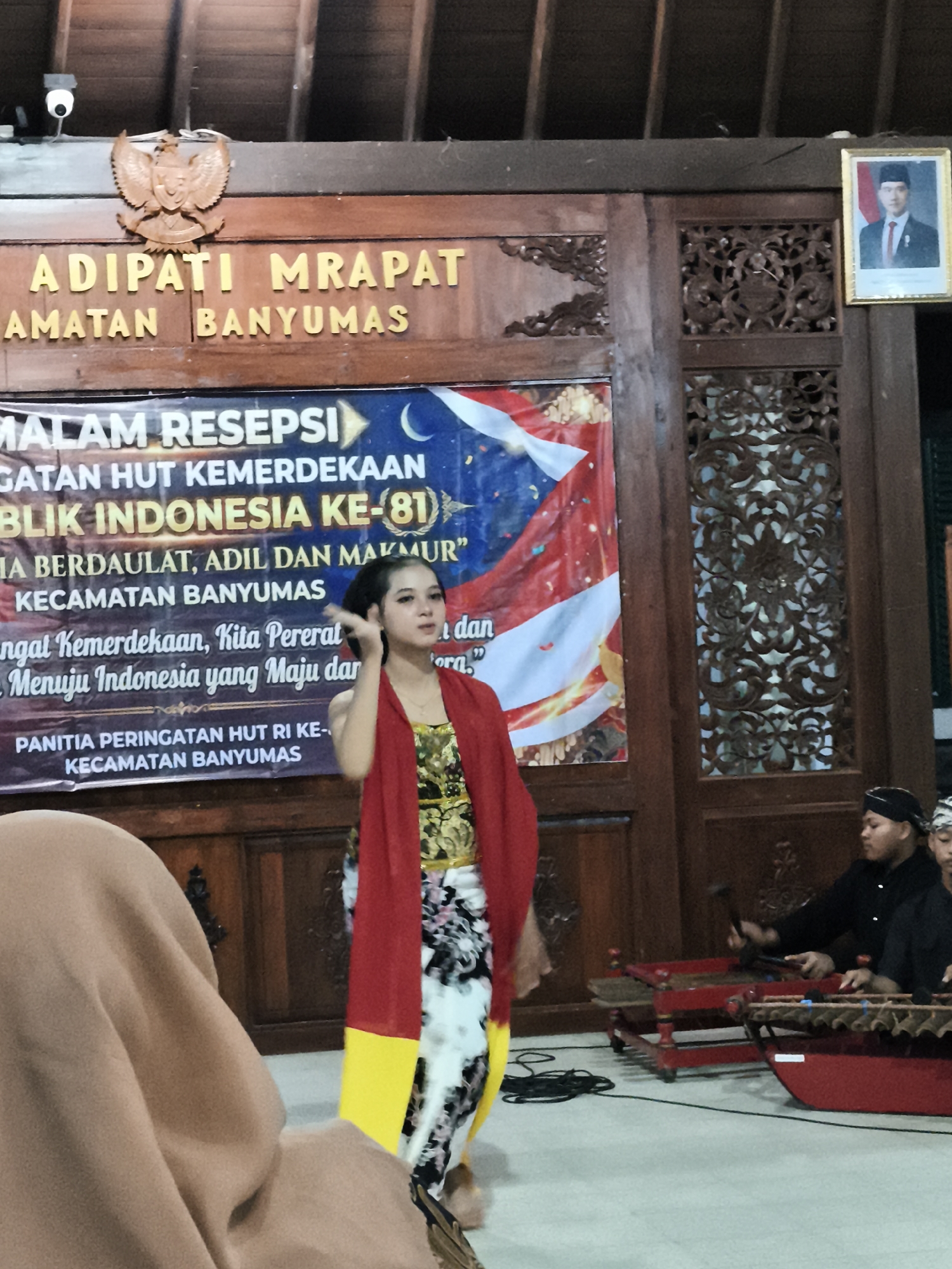 A young woman in traditional Indonesian attire, featuring a red scarf, a gold and green patterned top, and a black and white skirt with yellow accents, stands performing on a stage. She is positioned in front of a banner that reads "MALAM RESEPSI GATAN HUT KEMERDEKAAN REPUBLIK INDONESIA KE-81" and "IA BERDAULAT, ADIL DAN MAKMUR" "KECAMATAN BANYUMAS" and "PANITIA PERINGATAN HUT RI KE- KECAMATAN BANYUMAS". To the right of the stage, a group of men in black traditional clothing are playing gamelan instruments. The background features wooden carvings and a framed portrait of a dignitary. The setting appears to be a cultural event or ceremony in Banyumas, Indonesia.