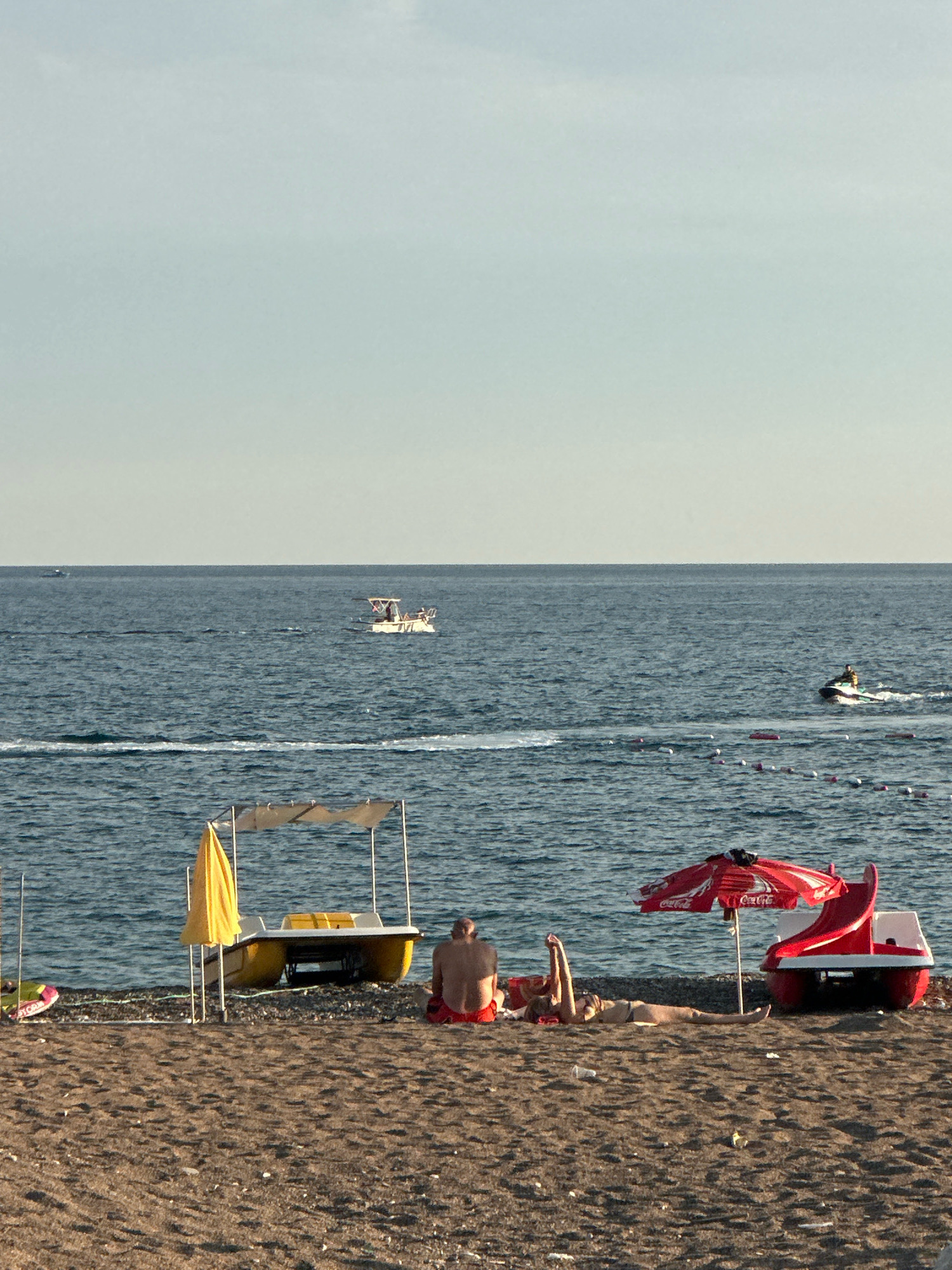 This image captures a serene beach scene in Boreti, Montenegro. The foreground is dominated by a sandy beach, littered with small pebbles and some debris. Two individuals are relaxing on the beach. A male, wearing red swim trunks, is seated with his back to the camera, facing the sea. Beside him, a female is lying down, seemingly sunbathing, with her arms raised above her head.
To their left, a yellow paddleboat with a canvas canopy is parked on the shore, with a yellow umbrella leaning against it. To their right, a red pedal boat with a slide, branded with "Coca-Cola," is positioned on the sand.
The middle ground features the calm, blue sea, with gentle waves lapping at the shore. Several buoys mark a swimming area. In the distance, a white boat with people on board cruises across the water, and a jet skier is visible further out, leaving a wake behind them. The horizon is a clear, pale blue sky, indicating fair weather. The overall impression is one of a peaceful, leisurely day at the beach.