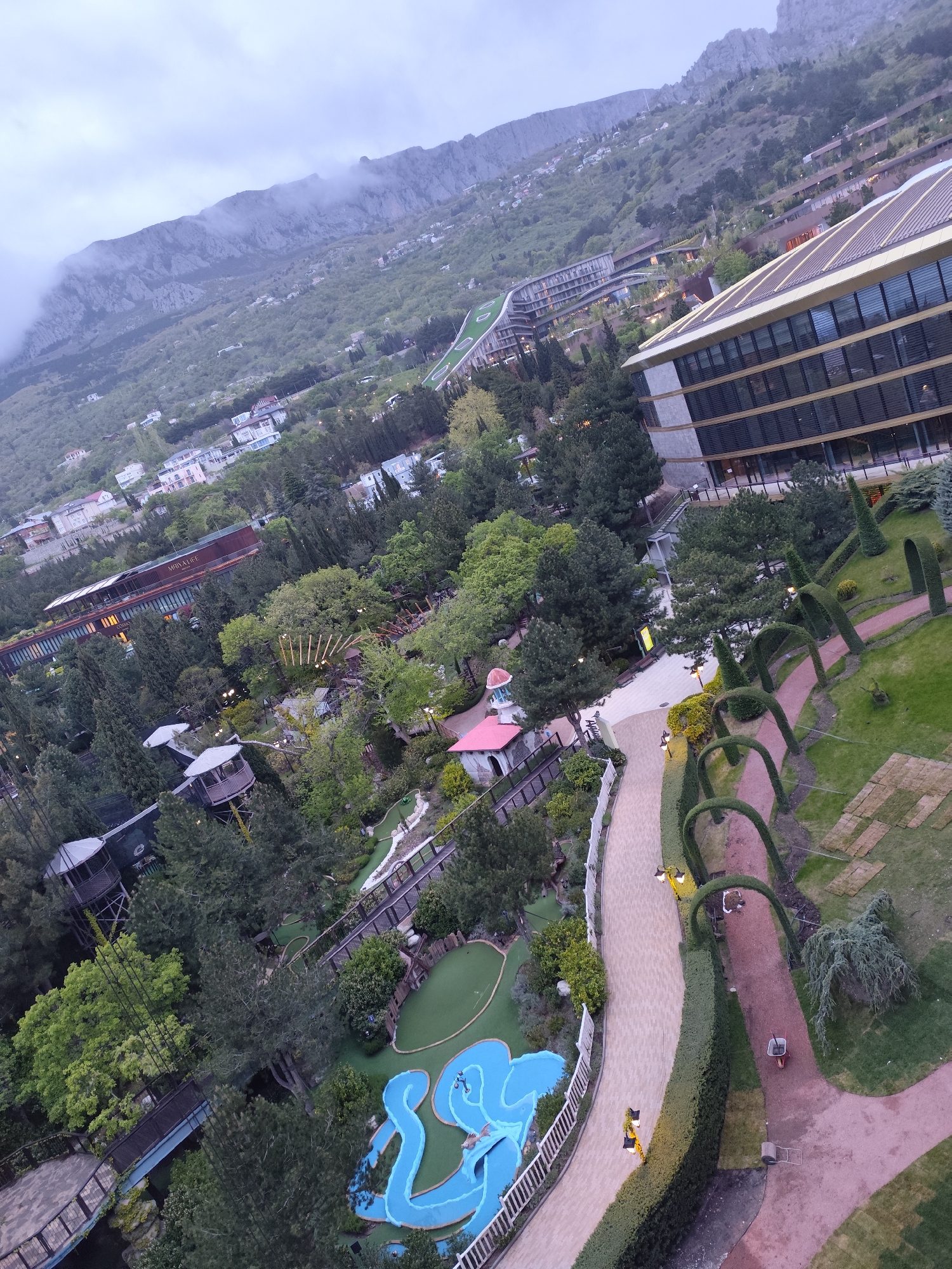 This is an aerial view in Симеизский поселковый совет, Ukraine, showing a landscape incorporating natural and man-made elements. A large, multi-story building with a modern design is situated on the right. Adjacent to it, a well-maintained garden features arched hedges lining a path. The center of the image contains a mini-golf course and treehouse structures amidst dense greenery. In the distance, a mountainous backdrop is partially obscured by clouds, with scattered buildings nestled on the slopes. The scene includes people walking on paths and structures that are part of this park or resort.