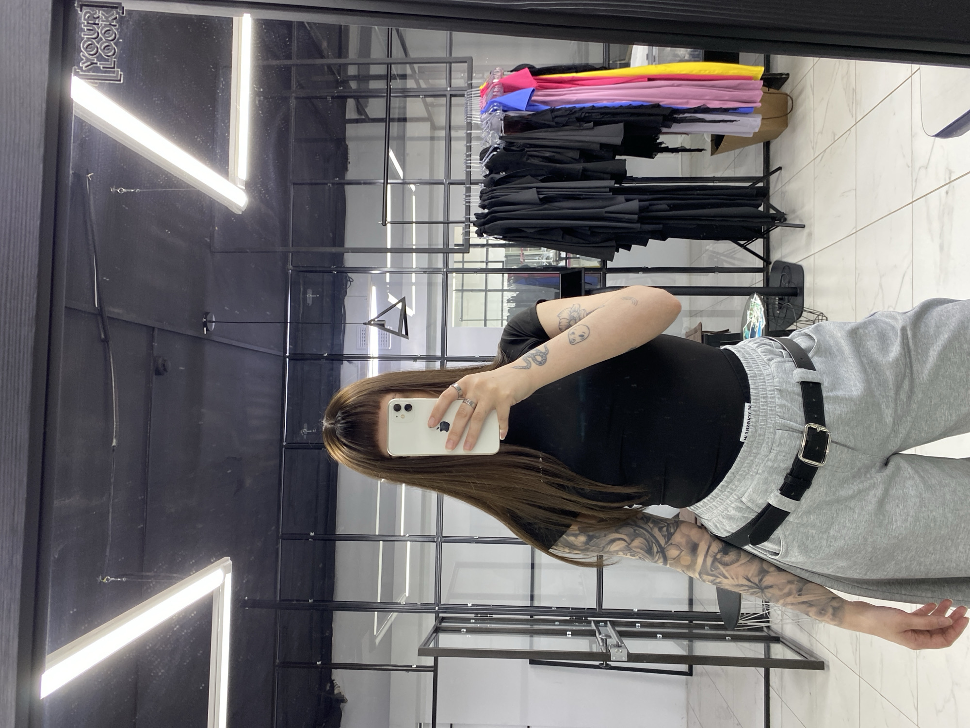 A woman is taking a selfie in what appears to be a retail clothing store, possibly in Votkinsk, Russia. She is holding a white smartphone in front of her face. The woman is wearing a black shirt and grey sweatpants, with a black belt cinched around her waist. Her arm is visibly tattooed. The background shows racks of clothing, and the store has a black interior with tiled flooring. There are rectangular lights on the ceiling. A mirror is visible, creating a reflection of the scene.