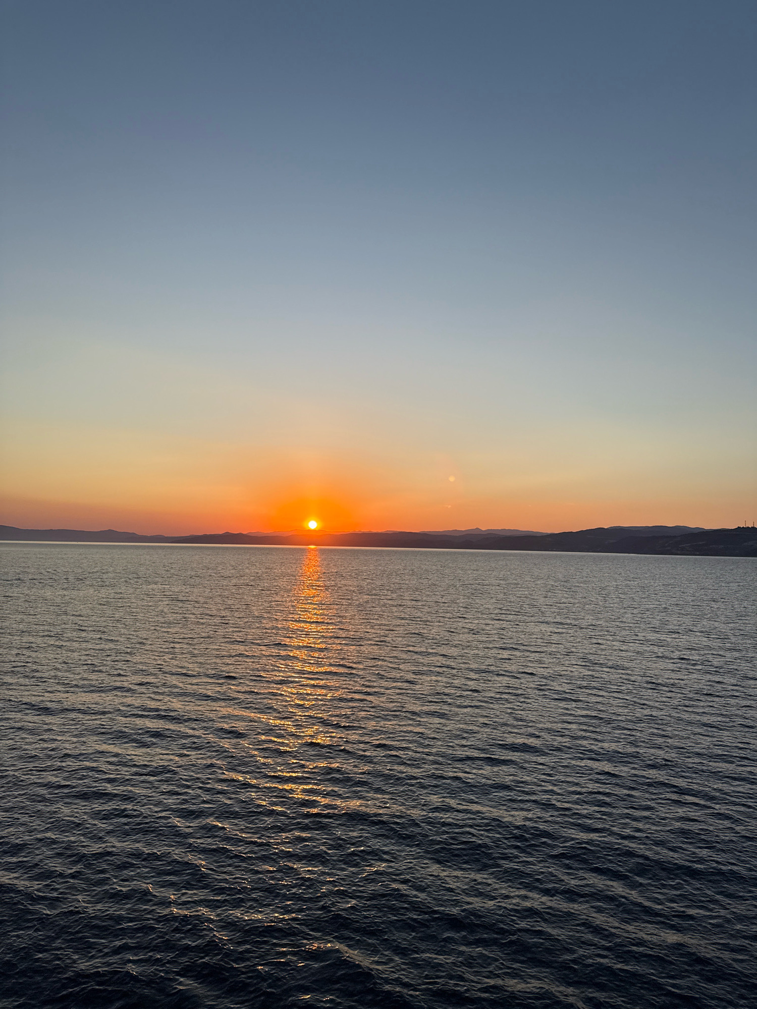 An outdoor, vertical, full shot captures a tranquil sunset over the Aegean Sea, as viewed from the coast of Ammouliani, Greece. The sun, a bright orange orb, sits low on the horizon, casting a shimmering golden reflection across the rippling, deep blue water. The sky transitions from a pale blue at the top to a warm gradient of yellow, orange, and pink near the horizon. Distant, silhouetted hills form a dark line against the colorful sky. The water appears calm with gentle waves, and there is no visible human presence or activity in the scene.