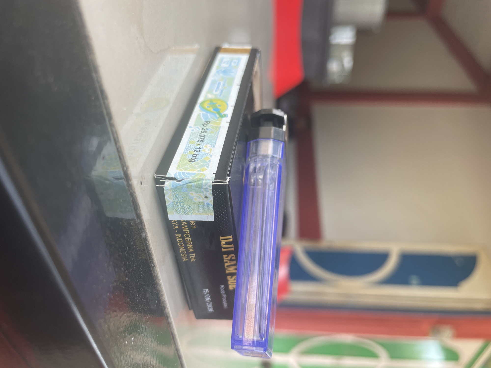A transparent blue cigarette lighter rests on top of a black cigarette pack. The cigarette pack has a holographic band around its side with text that reads "Rp 26.075 / 12 btg" and "DJI SAM SOE". The background is blurred, showing parts of a building with red, blue, and green accents, suggesting an outdoor setting. The scene appears to be an exterior shot in Muliengmanyang, Indonesia.
