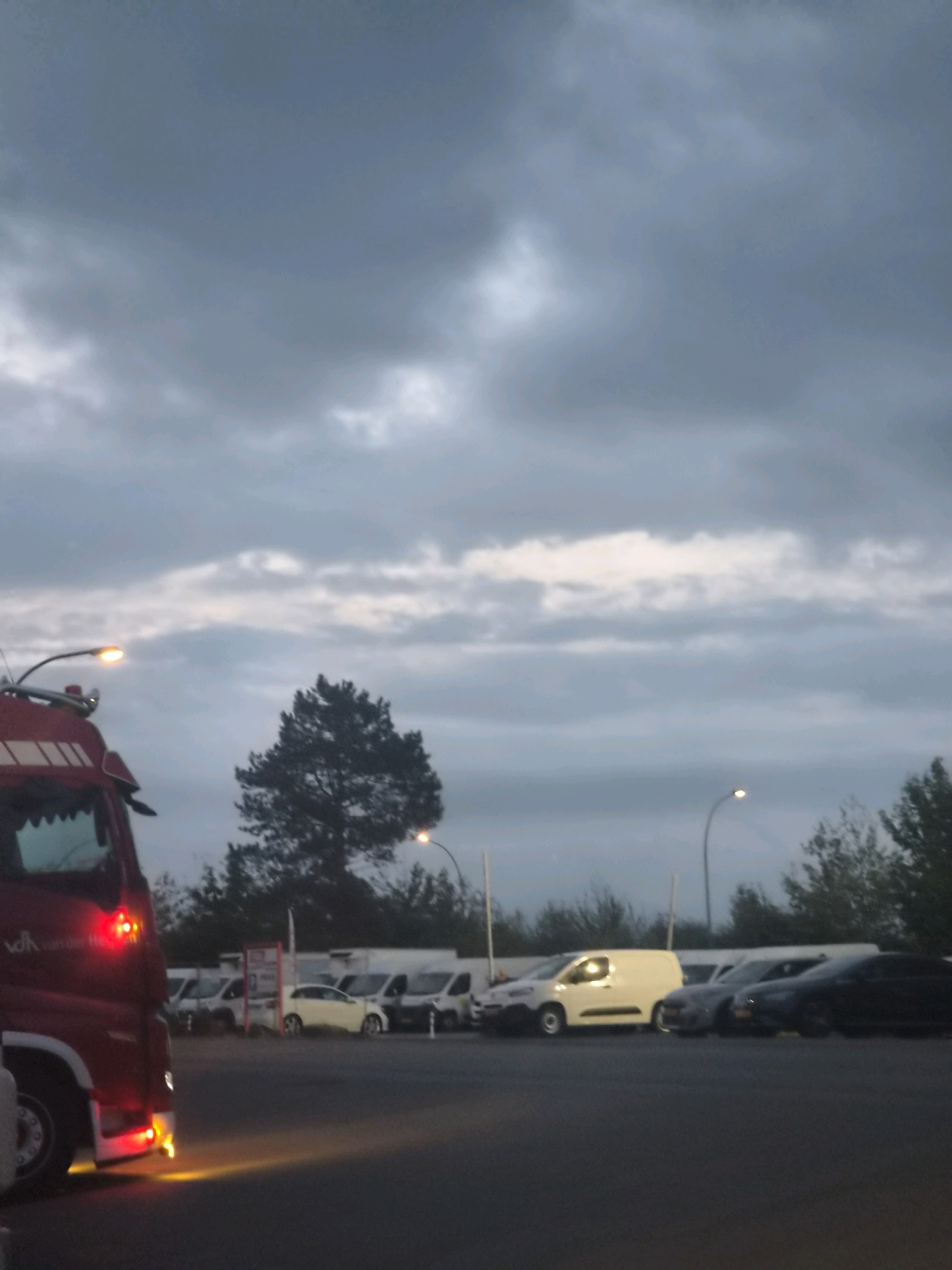 This image shows a parking lot in Leudelange, Luxembourg, under a cloudy twilight sky. In the foreground, a red truck is parked, with its headlights and hazard lights illuminated. The truck has visible branding on its side, which appears to be text. Behind the truck, several other vehicles are parked, including white vans and passenger cars. A single tall tree is visible in the middle ground, and streetlights are present throughout the scene. The overall lighting suggests it is either early morning or late evening.