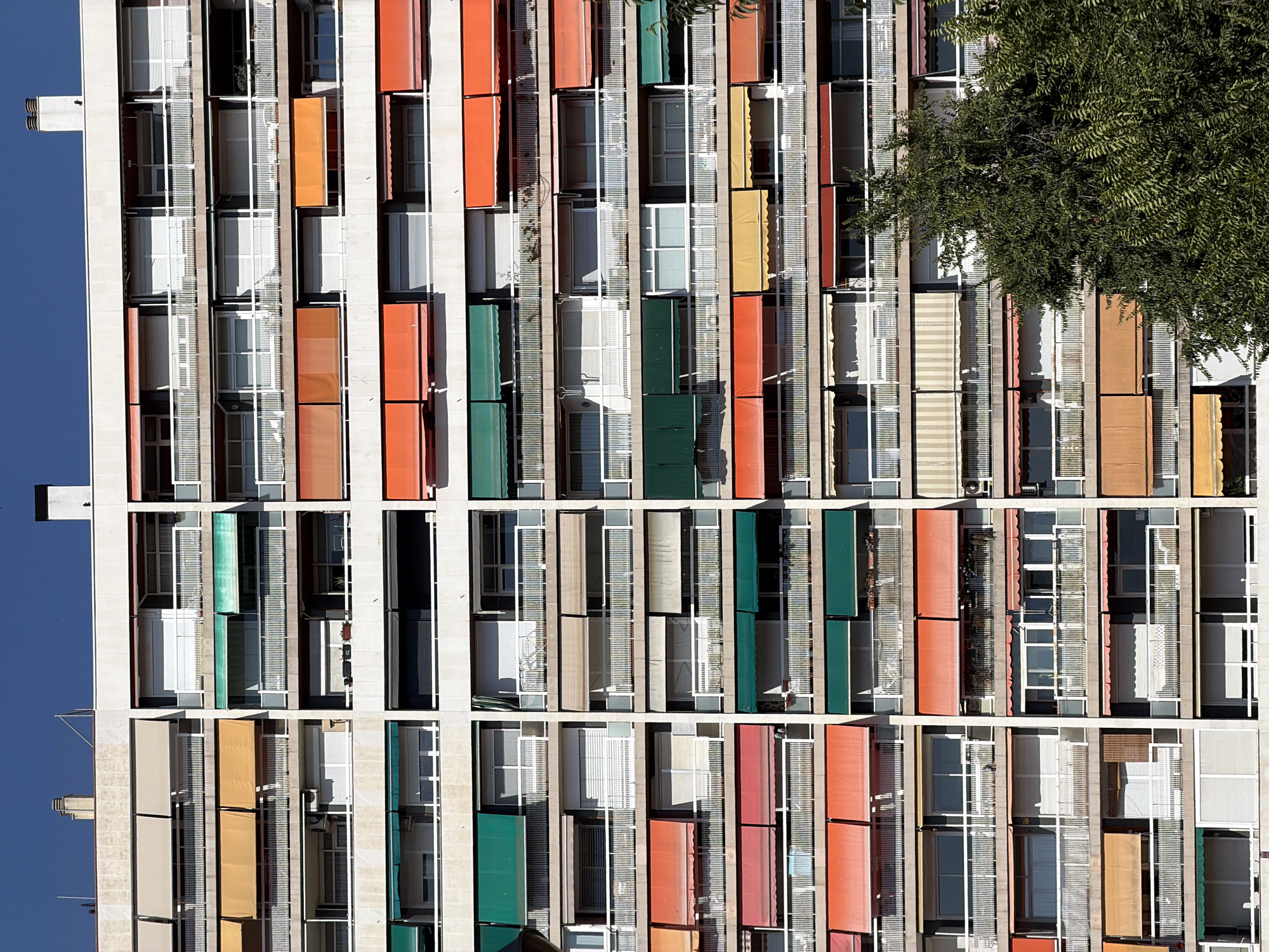 This is a high-angle, vertical shot of a colorful apartment building facade in Barcelona, Spain. The building is characterized by a repeating pattern of open and closed windows, balconies, and colorful awnings or screens. The architectural style appears modern, with a grid-like structure.
The scene is bathed in sunlight, suggesting a clear day and likely daytime, given the bright lighting and shadows. The building's exterior is a mosaic of different colored elements, including vibrant orange, muted yellow, deep green, and red, interspersed with white grates and grey or beige building material. Many of the balconies have large glass windows, some with white blinds or screens. The repeated vertical elements suggest multiple floors and apartments stacked one above the other.
On the right side of the image, the dense foliage of a large tree partially obscures the building, casting shadows on the facade. This suggests an urban setting with green spaces integrated into or adjacent to residential areas. The sky, visible on the far left and bottom, is a clear, deep blue, further indicating good weather. There are no people visible in the frame, and the focus is entirely on the building's architecture and its visual interplay of color and form. The overall impression is one of a vibrant and dynamic urban environment.