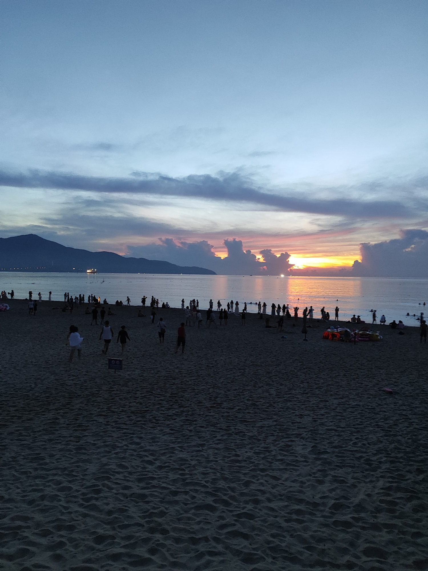 This is a wide-angle, low-angle shot of My Khe Beach in Da Nang, Vietnam, at dusk. The sky is a gradient of pale blue to soft gray, with streaks of pink and orange near the horizon where the sun is setting behind a range of silhouetted mountains. Scattered clouds are present in the sky, some tinged with the sunset colors.
The sea is calm, reflecting the sky's colors, and small boats or ships are visible in the distance on the water. The foreground is dominated by a sandy beach with visible ripples from the tide or wind. Numerous people, silhouetted against the water and sky, are spread across the beach. Some are walking, others appear to be standing or sitting. A few are in the shallow water.
In the middle ground, near the water's edge, a group of people are gathered, and a few more are further down the beach. Towards the right side of the image, closer to the viewer, several colorful objects that appear to be beach toys or inflatables are piled on the sand, along with some orange life vests. A small blue sign with white symbols is planted in the sand in the mid-foreground. The overall impression is a serene evening scene with a moderate number of beachgoers enjoying the end of the day.