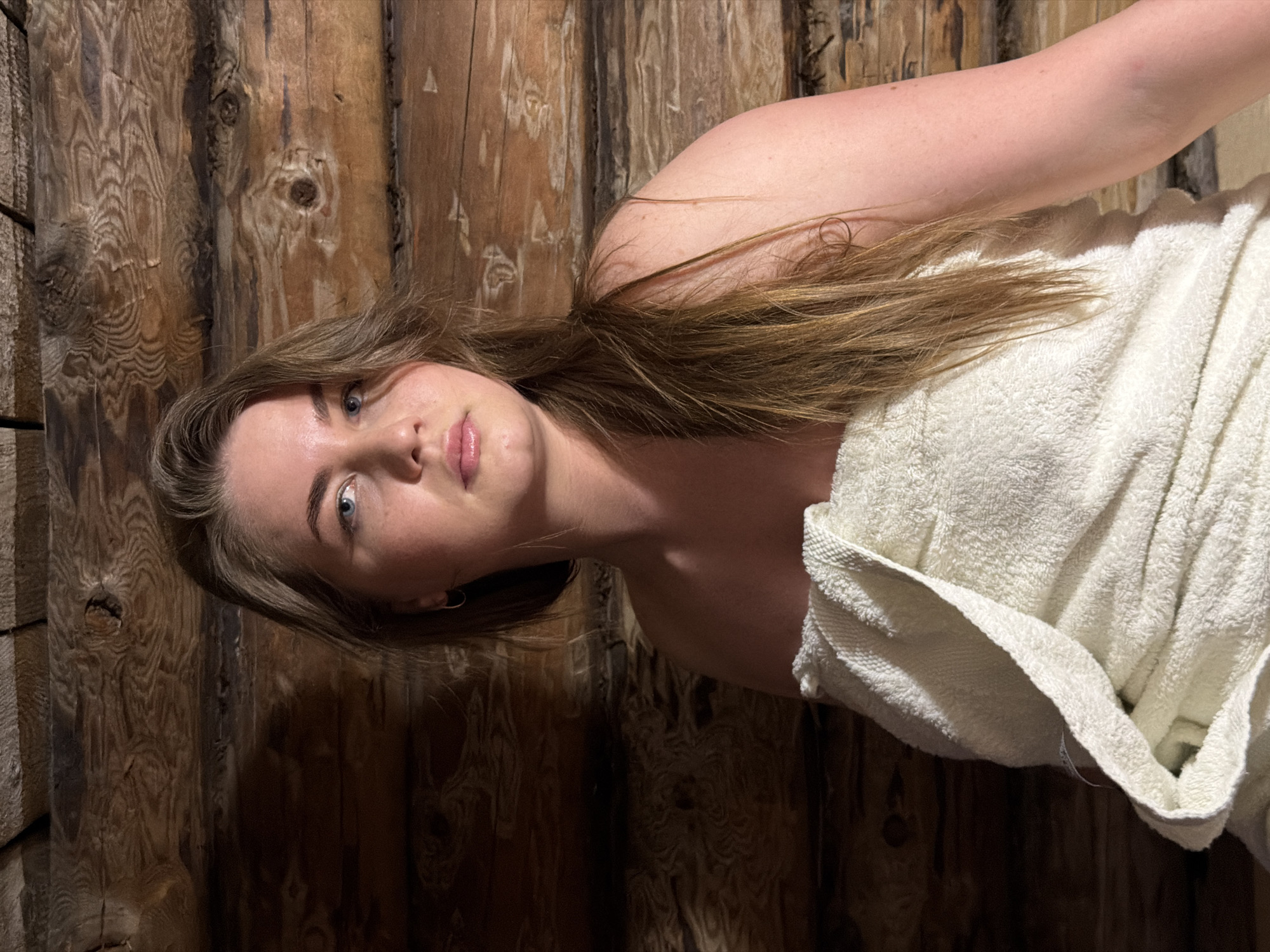 A woman with light skin and light brown hair wrapped in a white towel stands against a wall made of brown wooden logs in Уральское, Russia.