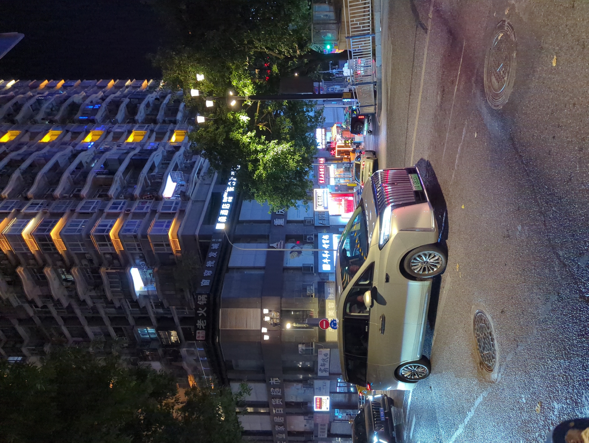 In Yuzhong District, China, at night, a modern, multi-story building with illuminated windows dominates the background, its facade featuring decorative orange lighting. In the foreground, a silver minivan is parked on a wet city street, its headlights illuminated. To the left of the minivan, the rear of a dark-colored car is visible, also with its headlights on. The street shows signs of recent rain, with reflections on the asphalt. Several streetlights are visible, illuminating the scene. Signs with Chinese characters are displayed on the buildings, suggesting commercial establishments.
