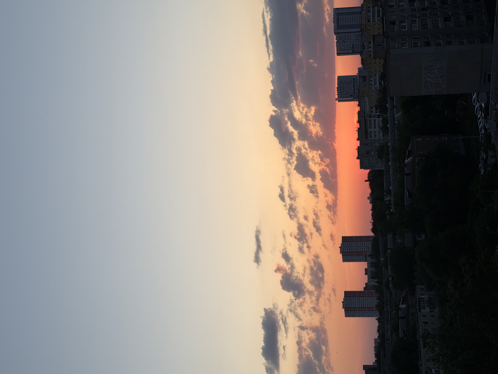 The image shows a cityscape at sunset. The sky transitions from a light blue on the left to orange and pink near the center and right, with scattered clouds tinged with orange and gray. Buildings, including high-rise apartment blocks, are silhouetted against the colorful sky on the right side of the image. The city's vegetation is also silhouetted. The location is Unknown, Unknown.