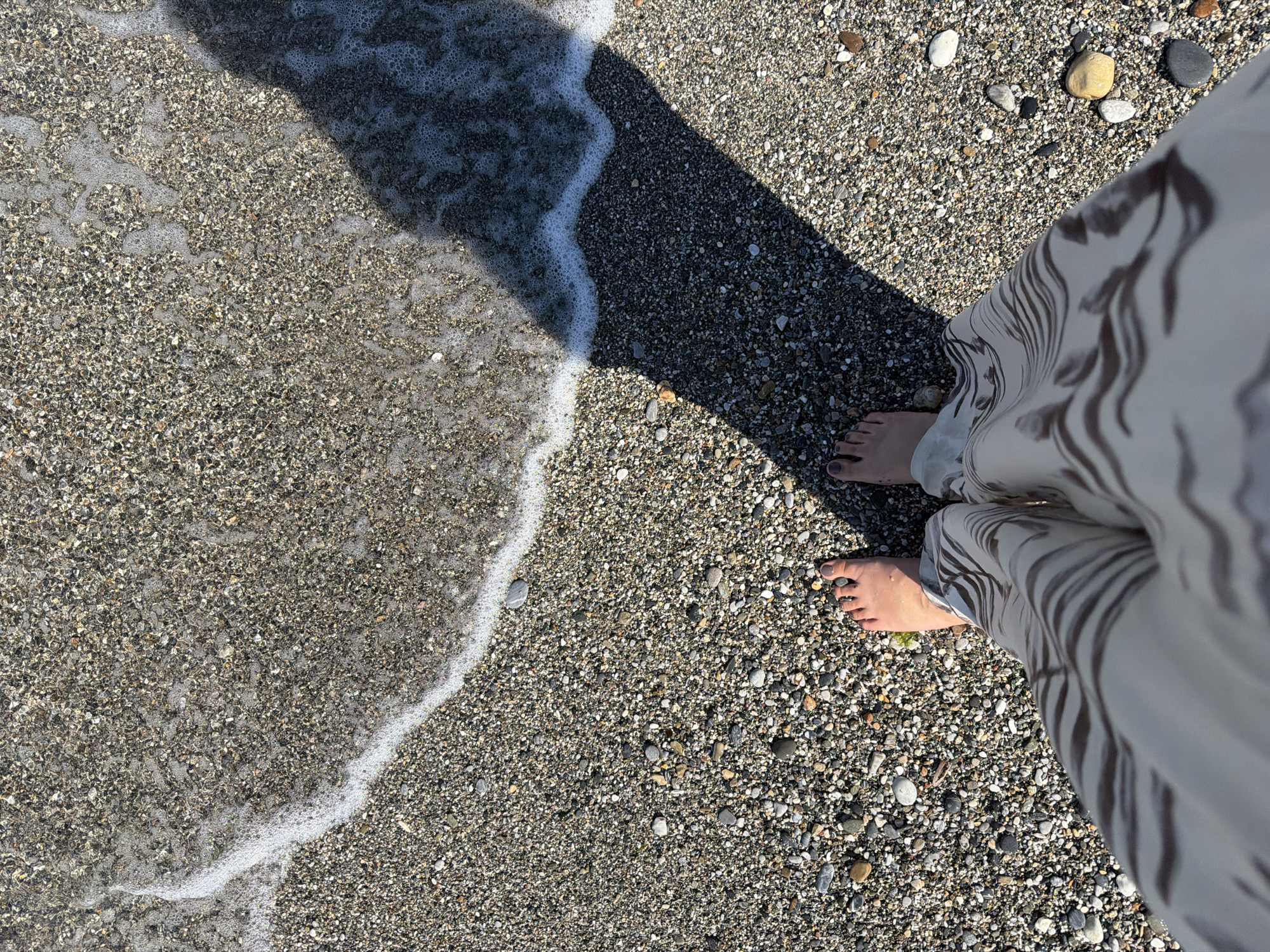The image shows a person's feet standing on a pebble beach, with the edge of the water lapping at the shore. The pebbles are small and varied in color, ranging from light gray to dark brown. A white foamy wave is visible, partially covering the pebbles. A shadow is cast diagonally across the beach, suggesting a bright sunny day. The person is wearing light-colored pants with a dark, abstract pattern. The scene appears to be on a beach in Alanya, Turkey.