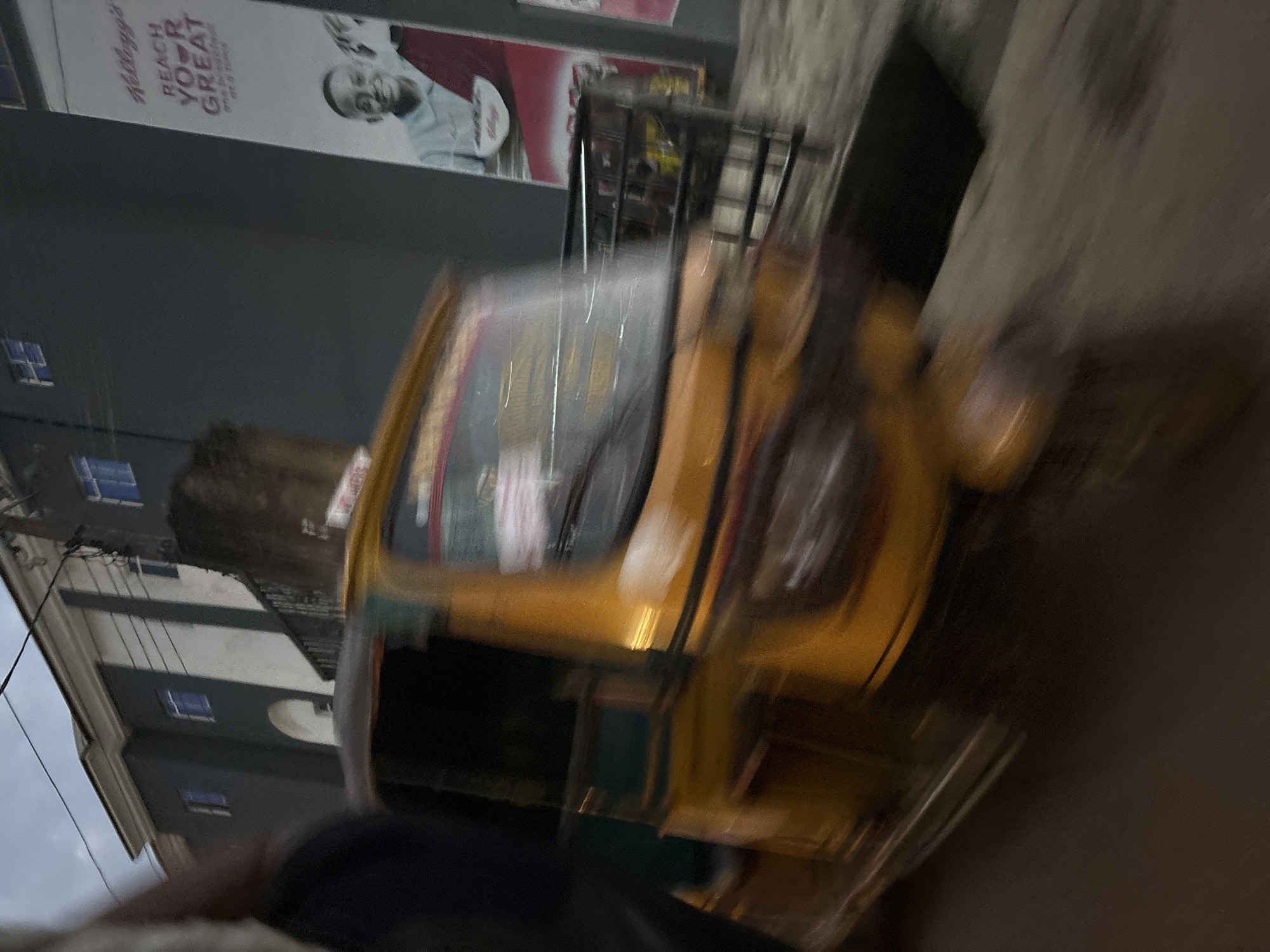 This is a motion-blurred photograph taken at night in the city of Onitsha, Nigeria. The main subject is a yellow auto-rickshaw, also known as a "Keke Napep," that is in motion. The background features a dark, modern building with white accents and windows, and a large advertisement for Kellogg's cereal with the slogan "REACH YOUR GREAT." A portion of another building is visible to the left, with a dark, blurry foreground element, possibly a person or another vehicle, obscuring part of the scene. The overall impression is one of urban movement and advertising.