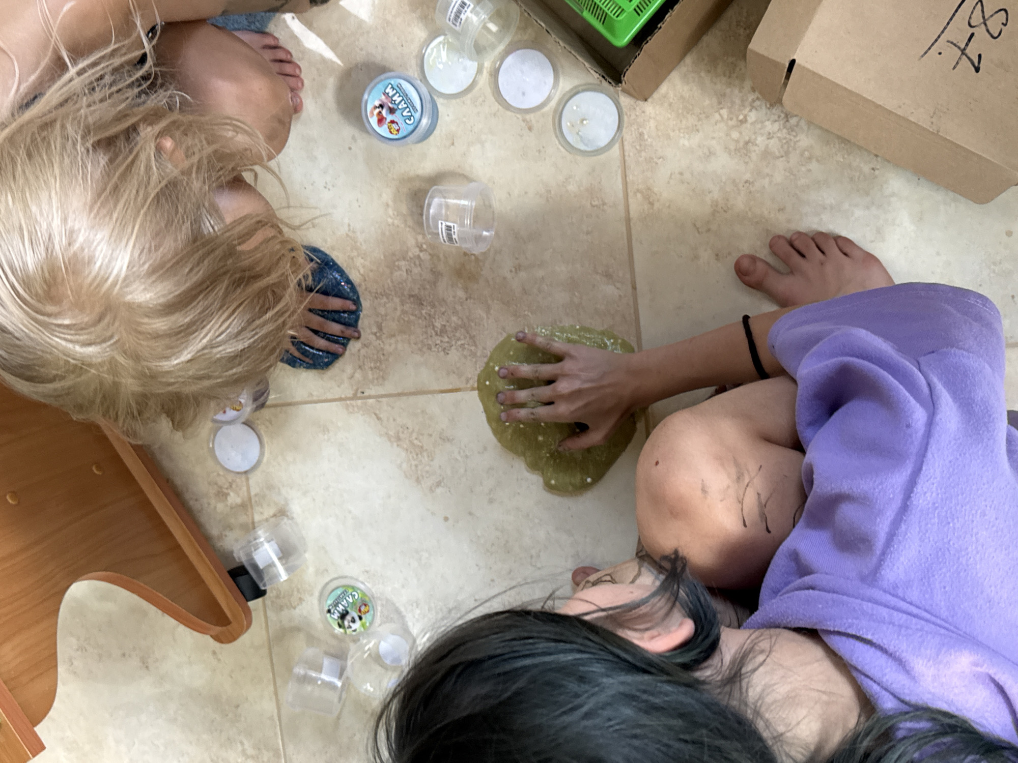 The image shows a top-down view of two children playing with slime on a tiled floor. One child, with blond hair, is leaning over a blue, glittery slime. The other child, with dark hair, is wearing a purple robe and is touching a light green slime. Several small, clear plastic containers, some with lids, are scattered around the slime. A wooden furniture piece is visible in the upper left corner. The environment appears to be an indoor residential setting. The location is not identifiable within the image.