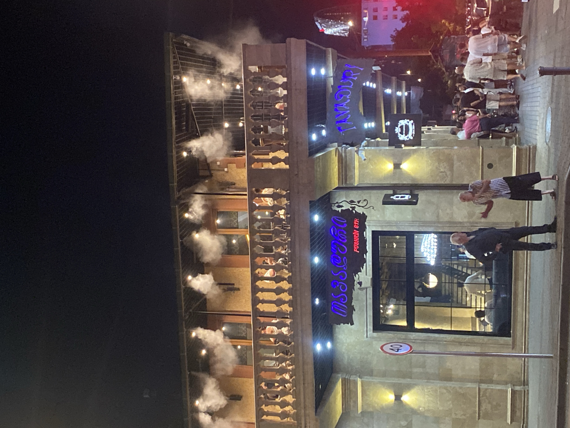 This is a nighttime street-level, medium-distance shot of a building with a balcony and multiple floors in Batumi, Georgia. The building's facade is made of light-colored stone, and there are ornate railings on the balcony. Various signs are illuminated with neon and traditional lighting. One sign above an entrance reads "TAVADURI" in blue neon letters. There is also a sign in Georgian script. Several people are visible on the street in front of the building, some standing in groups and others walking. One man in the foreground is gesturing with his hands. The upper floors of the building have a covered walkway or terrace with decorative lights and mist emanating from it. In the background, other buildings, including a tall, illuminated skyscraper, are visible. A speed limit sign with "40" is posted on the left side of the street. The overall scene suggests a vibrant nightlife or dining area.
