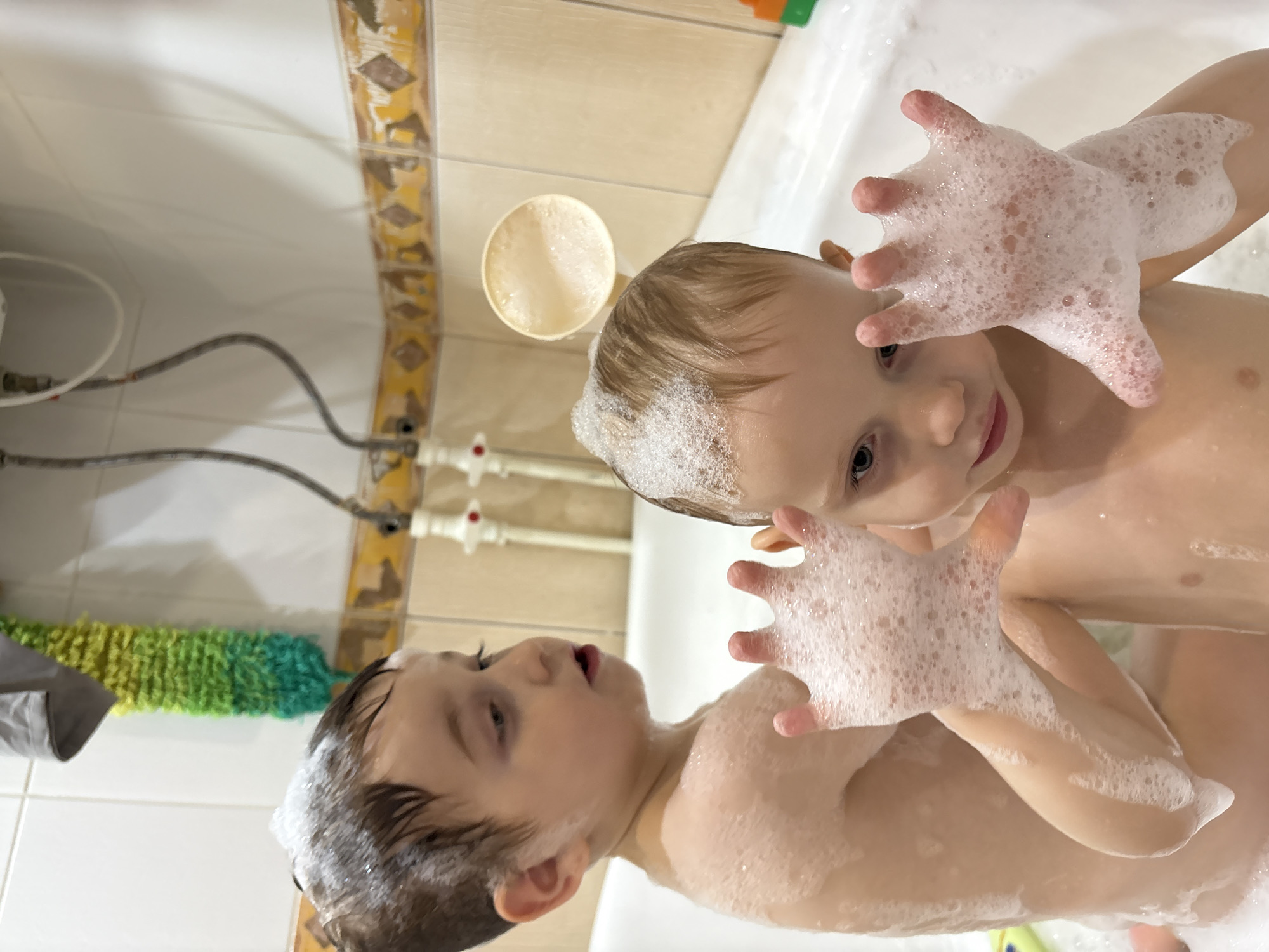Two young children are in a bathtub filled with water and covered in soap bubbles. The child on the right has blonde hair and is looking downward with a slight smile. Their hands are raised, covered in bubbles. The child on the left has brown hair and is looking to the right with an open mouth. Their hands are similarly raised and covered in bubbles. The location appears to be a bathroom, with tiled walls and visible plumbing fixtures. A green and yellow cleaning brush is hanging on the wall. A cup with foam is on the bathtub rim. The location is in the city of Chaikovsky, Russia.