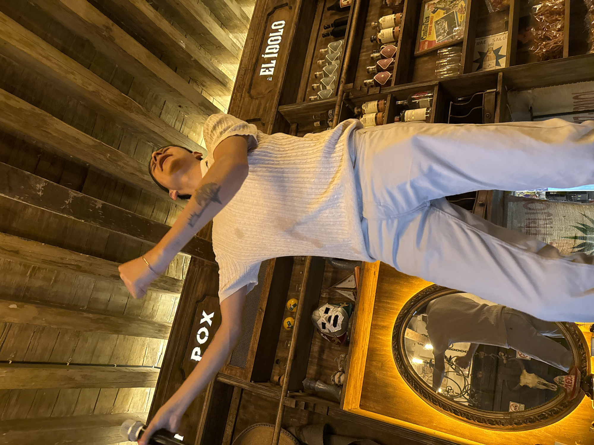 A man is holding a microphone in what appears to be a restaurant or bar setting in the city of Puebla, Mexico. The building’s interior is constructed with wood. The man is wearing a white top and light-colored pants. The shelves behind him are lined with wine bottles, assorted decorative objects, and vintage advertisements. A mirror is visible, reflecting the man and the restaurant decor. The name “EL IDOLO” is displayed on a vertical wooden board.