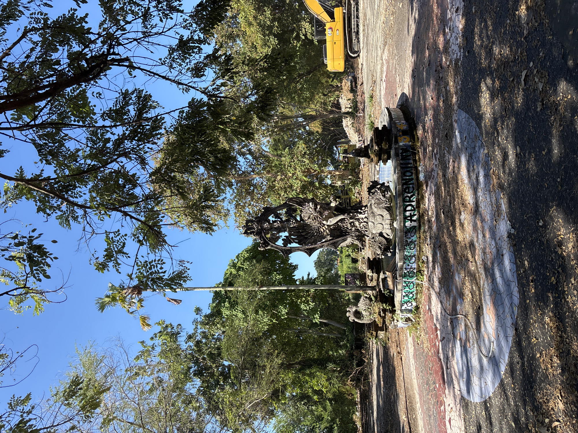 The image shows a stone fountain covered in graffiti in a park in Denpasar, Indonesia. The fountain is decorated with carved dragons. A yellow excavator is visible in the background, and trees and a blue sky are visible above the fountain. The ground around the fountain is covered in leaves and debris.