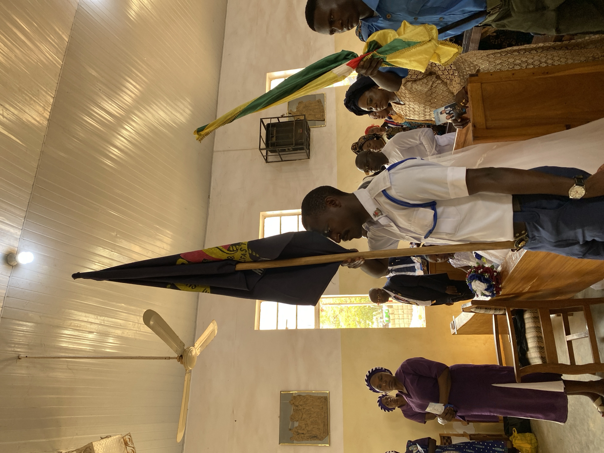 The image shows an indoor scene in Kuru Dungus, Nigeria, presumably a church or similar building, evident from the pews and religious articles. A group of people, mostly African, are present. A man in a white uniform with blue trim is holding a black flag with yellow and red accents. Another man holds a green, yellow, and red flag. Other people are seated, appearing to be observing or participating in a ceremony. The room features white walls, a ceiling fan, and a few windows.