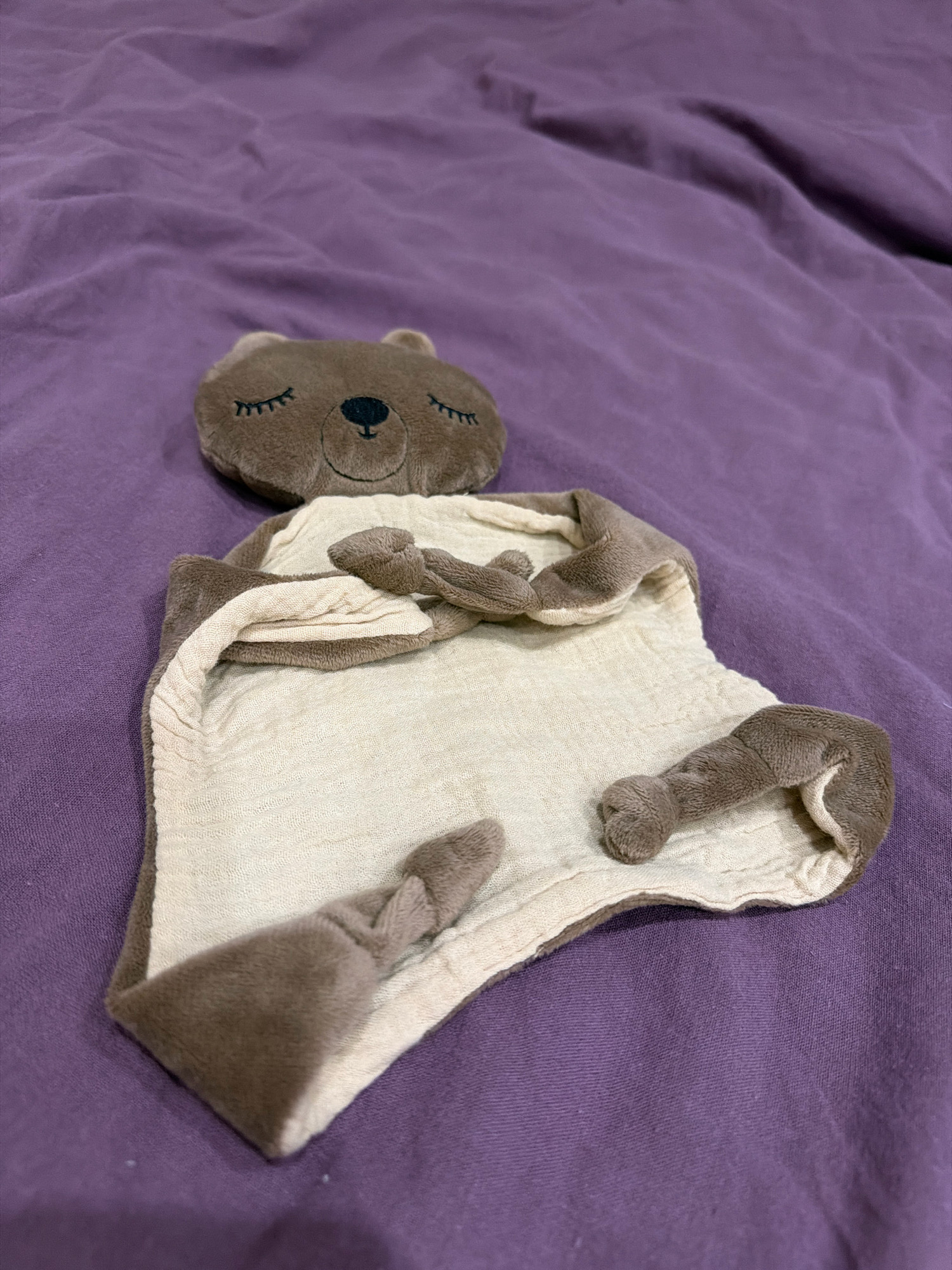 A close-up shot shows a brown teddy bear security blanket laid flat on a wrinkled purple fabric. The bear's head is at the top, with its eyes closed and a gentle smile. The blanket itself is a light beige color with textured white fabric underneath, and it is folded in places. The bear's arms and legs, made of brown fuzzy material, are extended outwards. The purple fabric beneath is a solid color and appears to be a bedsheet or similar material. The lighting is soft, and the image is taken from a slightly elevated angle. There are no people or animals present in the image. The location is not specified in the provided context.