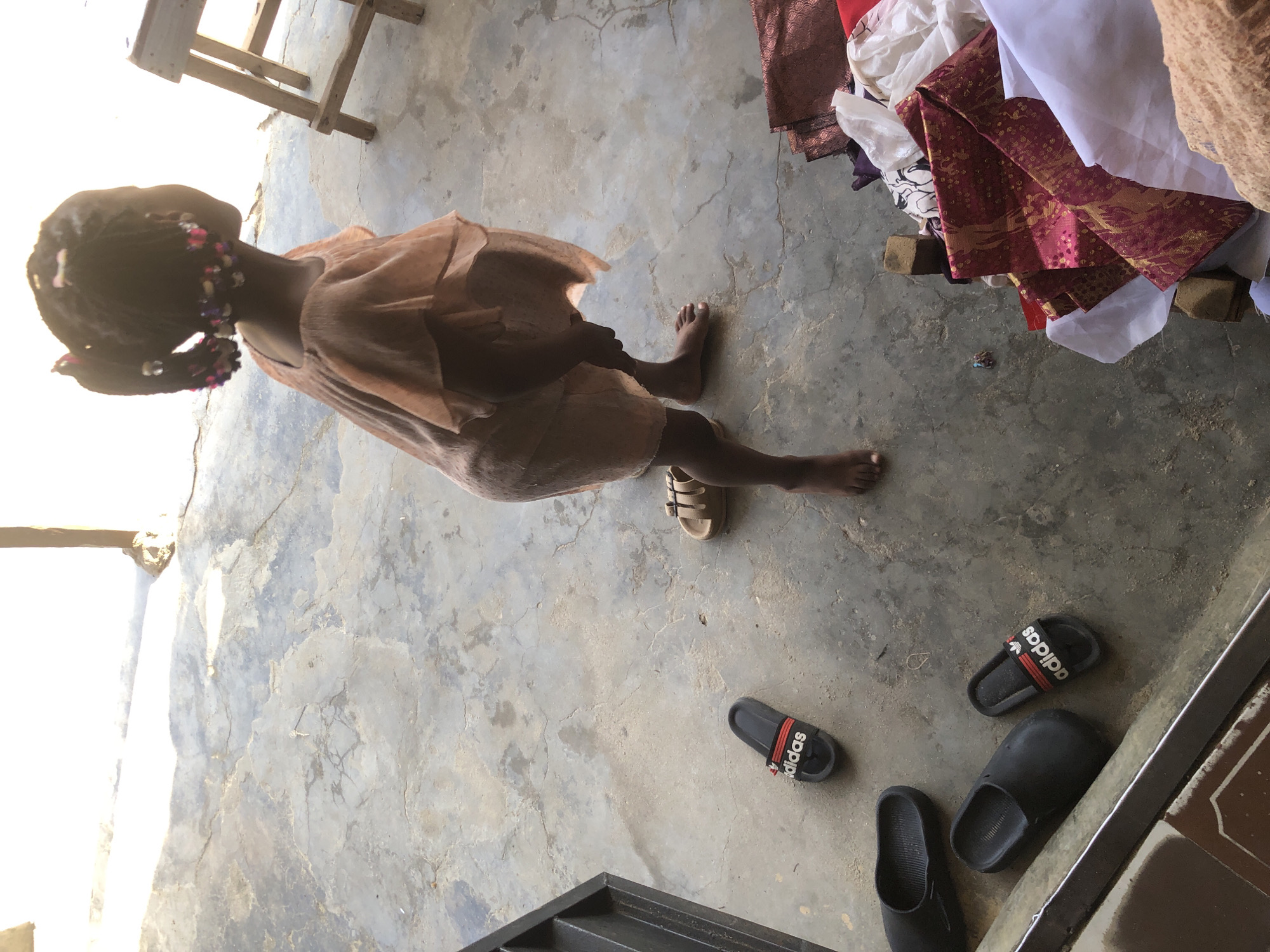 The media file shows a young Black child from a top-down perspective, standing on a concrete floor. The child is wearing a loose, light brown dress and has dark hair styled in braids adorned with colorful beads. The child's back is to the camera, and they are facing away from a pile of colorful fabrics, some with intricate patterns in red, gold, and white. To the left of the child, a wooden stool is visible, and further to the left, a white wall or structure.
Scattered on the concrete floor are several pairs of sandals. Close to the child's feet, there are two pairs of black Adidas slides with red and white stripes. There are also at least two other black slides and a pair of tan slide sandals.
The scene appears to be outdoors or in an open-air setting, possibly a workshop or a storage area given the presence of fabrics and building materials. The lighting suggests it is daytime, with sunlight casting shadows. The concrete floor is weathered and cracked, indicating an outdoor or utilitarian space. The overall impression is one of simple, everyday life in a rural or semi-urban environment. There is no discernible emotional expression from the child. The visible text is "adidas" on the slides. The location is identified as Ado, Nigeria.