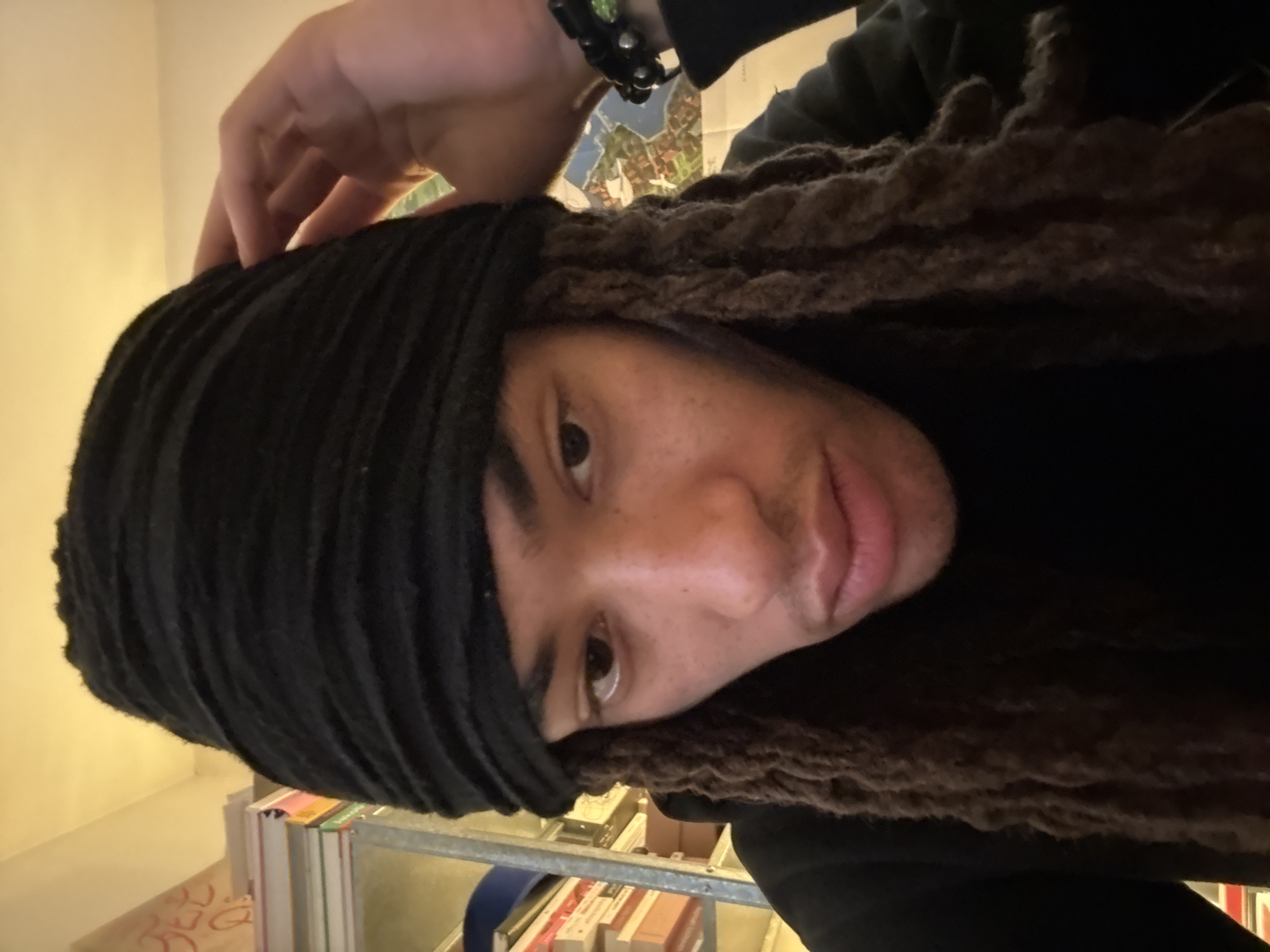The scene depicts a close-up, head-and-shoulders portrait of an individual within an indoor setting in Paris, France. The subject, appearing to be male, has medium-brown skin, dark brown dreadlocks, and wears a black, textured head covering over the forehead and ears. A black long-sleeved garment is also worn. The subject's left hand is raised, with fingers resting on the side of the head covering. A black beaded bracelet featuring silver-colored studs and a single green accent bead is visible on the left wrist. The individual looks directly towards the camera with a neutral expression. The background, largely out of focus, reveals a light-colored wall. To the subject's left, a metal shelving unit holds multiple books and papers, above which faint red graffiti-style lettering is discernible on the wall. To the subject's right, a colorful, stylized map or poster is affixed to the wall. The environment is illuminated by a warm, artificial light source.