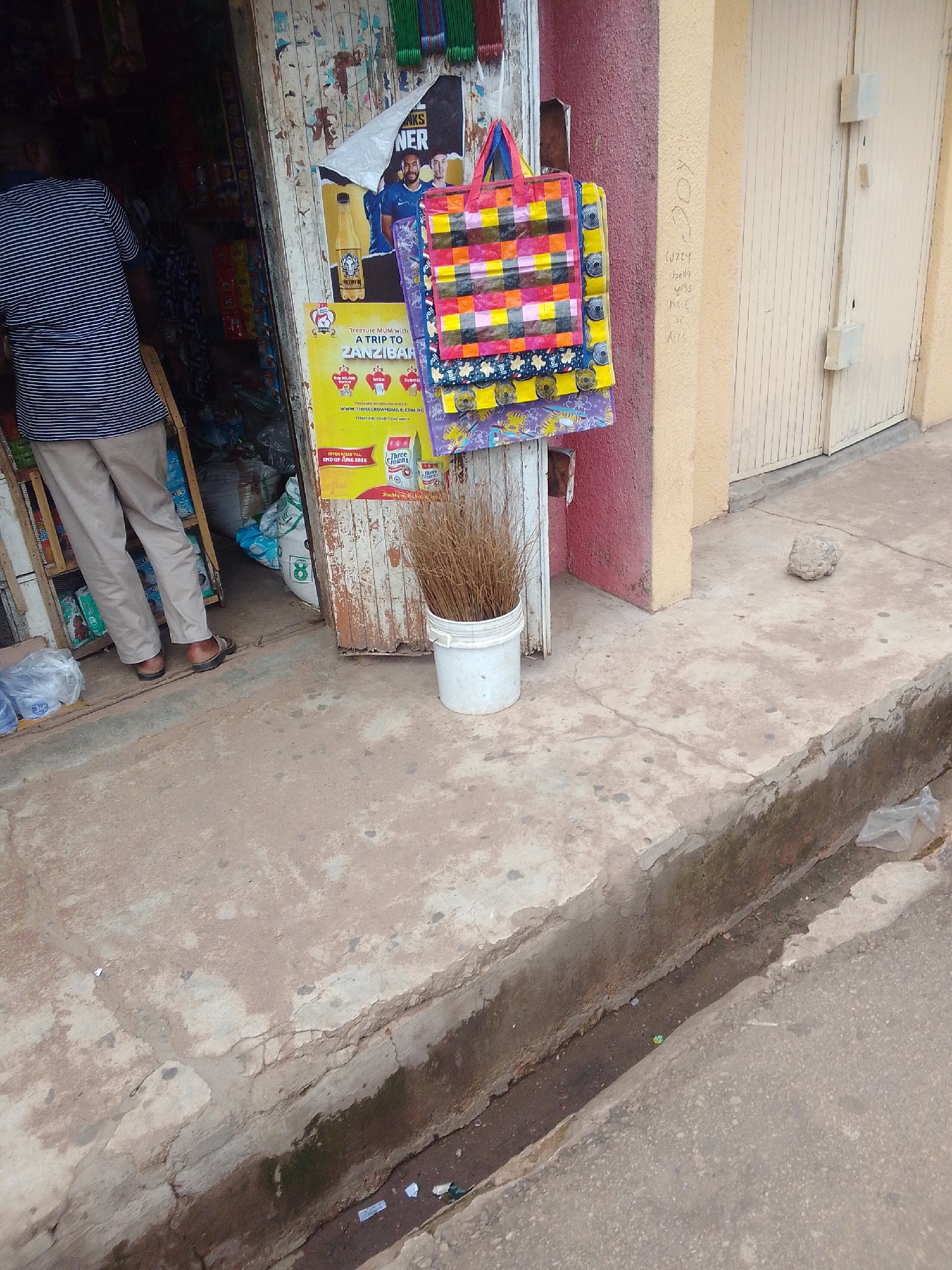 This image captures a street scene in Jos, Nigeria, focusing on the entrance of a small, modest shop.
In the left foreground, a person is visible from the back, standing within the shop's dark interior, facing deeper inside. They are wearing a blue and white horizontally striped t-shirt and light tan trousers, with dark sandals on their feet. Their posture suggests they are either an attendant working or a customer browsing.
The shop's entrance is framed by old, weathered wooden posts, with chipped paint. Several colorful, woven plastic bags, featuring checkerboard patterns in shades of pink, yellow, blue, and red, are hanging on display for sale on a hook attached to the wooden frame. Adjacent to the shop entrance, posters are affixed to the wall. A prominent yellow poster advertises "A TRIP TO ZANZIBAR" sponsored by "THREE CROWNS MILK," with a website address and a note indicating the offer is valid until "END OF JUNE 20XX." Above this, another poster shows two male figures, one appearing to be a football player, with partial text visible. Inside the shop, various goods, possibly sacks or packaged items, are dimly discernible.
To the right of the shop entrance, a white plastic bucket containing a bundle of dry, light brown grass or twigs stands on the sidewalk. The building wall immediately next to the shop is painted a reddish-pink, transitioning to a light yellow/cream wall at a corner. Faint, informal writing is visible on the yellow wall, including words like "Izzy" and "belly." A light-colored, vertically paneled door with two square security latches is set into this yellow wall.
The ground consists of a concrete sidewalk that appears worn, stained, and cracked. A small, irregularly shaped light-colored rock rests on the sidewalk further from the shop. A narrow, shallow drainage ditch runs along the edge of the sidewalk, containing some dirt, small pieces of litter, and a clear plastic bag. The ditch separates the sidewalk from a darker, coarser street surface, partially visible in the lower right corner. The overall impression is an active, weathered urban commercial setting.