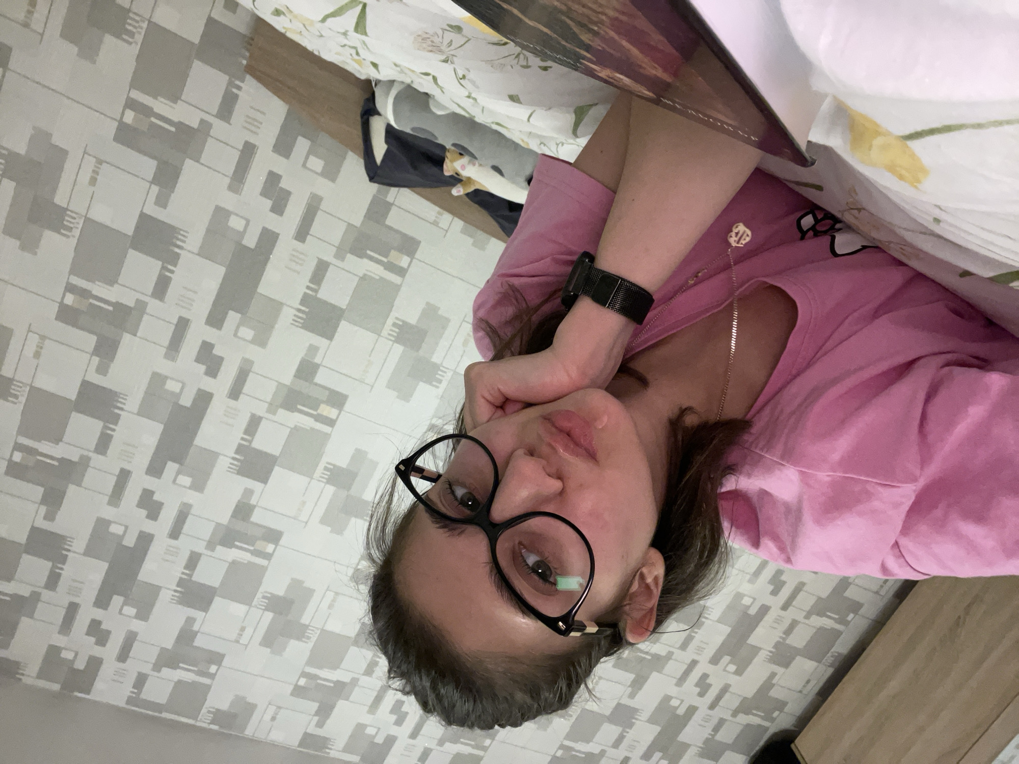A woman with dark hair, wearing glasses, a pink long-sleeved shirt, and a black watch, is lying down, propping her head up with her hand. She is indoors, presumably in a bedroom given the bed linens and patterned wallpaper. The image provides no information to determine the location is the city of Sofia, Bulgaria.