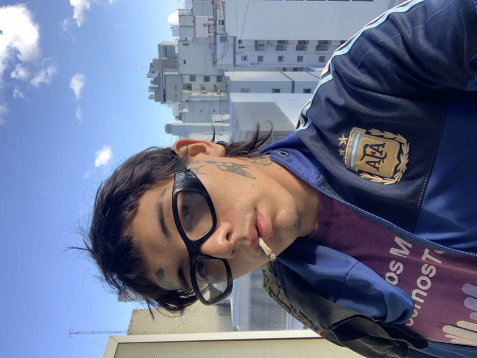 A man wearing large black glasses and an Argentina soccer jacket is holding a lit cigarette to his lips. He has tattoos on his face and neck. He is wearing a purple t-shirt with white text. The background shows buildings and a blue sky with clouds. The image is oriented vertically.