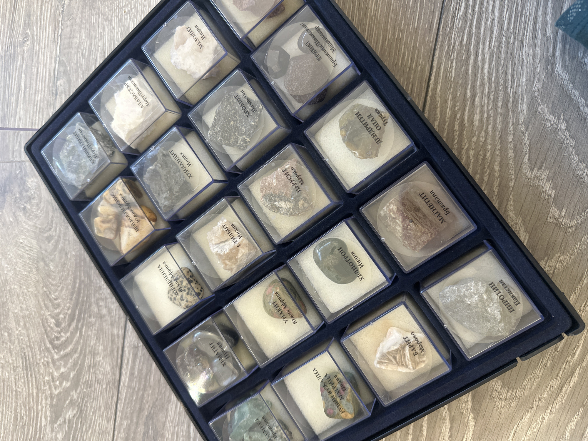 The image shows a collection of mineral specimens in a dark blue box with compartments. Each compartment contains a small sample of a mineral, enclosed in a clear plastic case with a label. The labels appear to be in Bulgarian, identifying the mineral and its origin (e.g., "Изумруд Бразилия" for Emerald from Brazil). Minerals visible include emerald, hematite, hornblende, ruby, unakite, stilbite, jasper, serpentine, heulandite, alabaster, barite, heliotrope, cerussite, chromite, mesolite, pyrrhotite, magnetite, dendrite opal, and praline. The box is placed on a surface with a wood grain pattern. The image does not suggest that the scene is located in Sofia, Bulgaria.