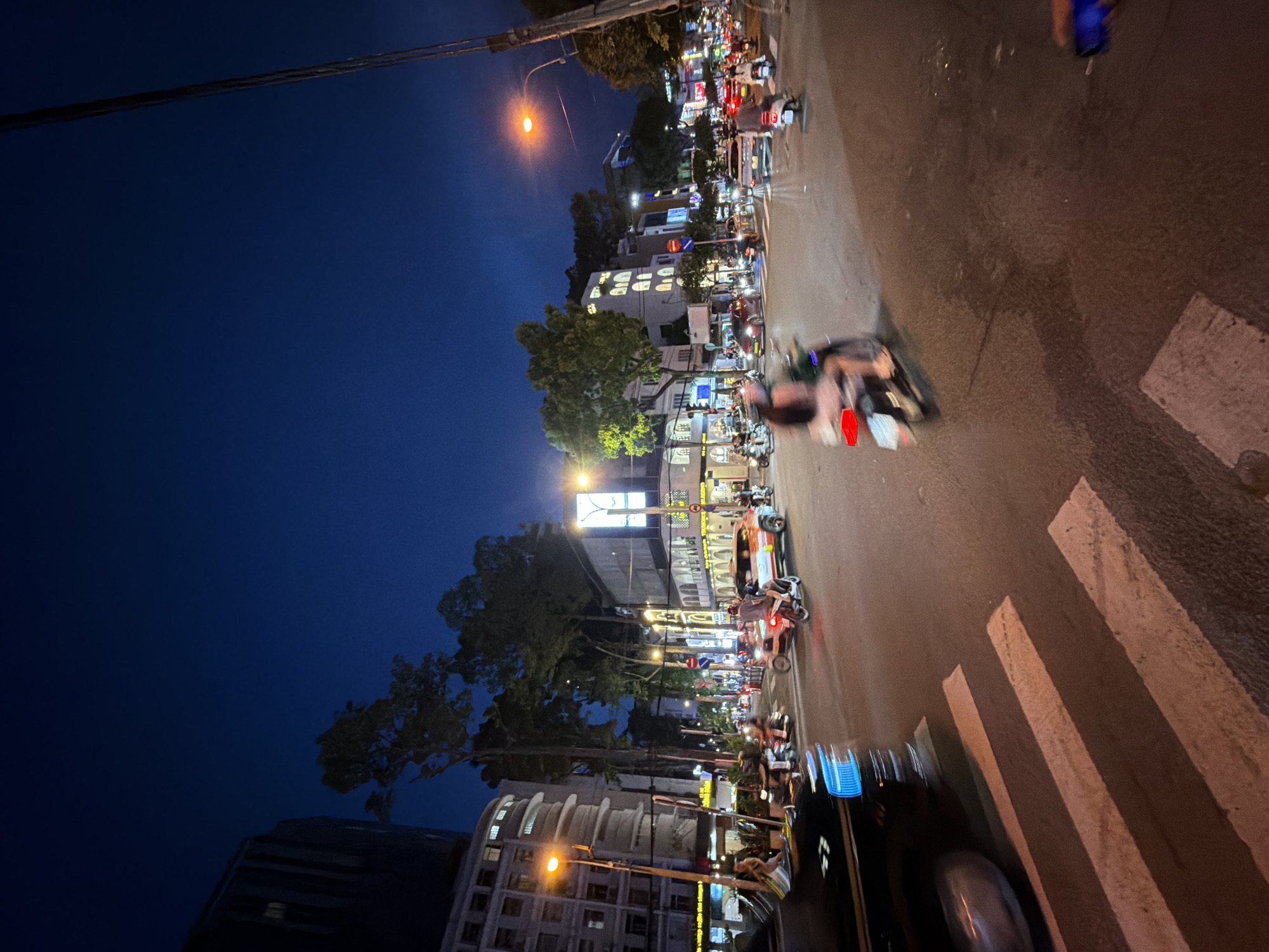 The image depicts a busy city intersection at dusk in Thủ Đức, Vietnam. The sky is a deep twilight blue, transitioning from night. Streetlights and illuminated signs from businesses cast a warm glow on the wet asphalt road, reflecting the lights of vehicles. Pedestrian crossings are visible in the foreground.
Numerous motorbikes, the primary mode of transportation in Vietnam, navigate the intersection. Some are in motion, creating motion blur, while others appear stationary. A few cars are also present, including a distinctive orange taxi with white lettering and a dark-colored SUV in the left foreground.
People are seen riding the motorbikes, some with passengers. A few individuals are also visible on the sidewalks, near shopfronts. The buildings lining the street showcase a mix of modern and older architectural styles, with illuminated windows and signs indicating commercial establishments. Trees with full foliage are interspersed between the buildings, their dark shapes contrasting with the bright lights. The overall impression is one of a vibrant, active urban environment at night, characteristic of a bustling Vietnamese city.