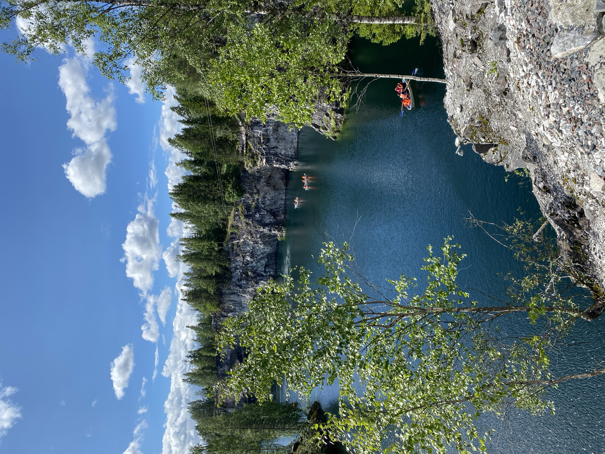 The image depicts the Marble Canyon in Ruskeala, Russia, on a sunny day. The canyon features a striking body of turquoise water surrounded by steep, rocky cliffs. A dense forest of green trees covers the tops of the cliffs and extends into the background. Several small boats, each with occupants wearing orange life vests, are visible on the water. One boat is closer to the foreground, with two individuals rowing. The sky is a clear blue, punctuated by fluffy white clouds. The overall scene conveys a sense of natural beauty and recreational activity in a unique geological setting.