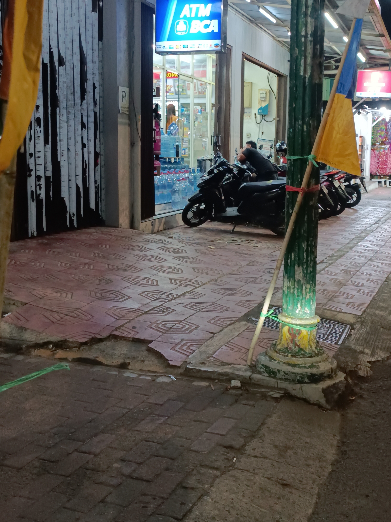 The image captures a street scene in Yogyakarta, Indonesia, likely at dusk or night, given the lighting. In the foreground, a textured, patterned tile pavement shows signs of wear and damage, with broken sections and exposed earth. A green plastic hose lies coiled on the ground to the left. A decorative, fluted lamppost, painted green and adorned with yellow and red at its base, stands prominently in the mid-ground. Tied to the lamppost with red and green rope is a bamboo pole, which in turn supports a yellow and blue flag. Behind the lamppost, several parked motorcycles are visible, with one person seated on a black scooter, facing away from the camera. Further back, a storefront with an illuminated blue "ATM BCA" sign is visible, suggesting a financial institution or a service area. Inside the store, rows of bottled water are stacked, and several individuals, including one with orange hair, are visible. The overall environment suggests a busy commercial or pedestrian area.