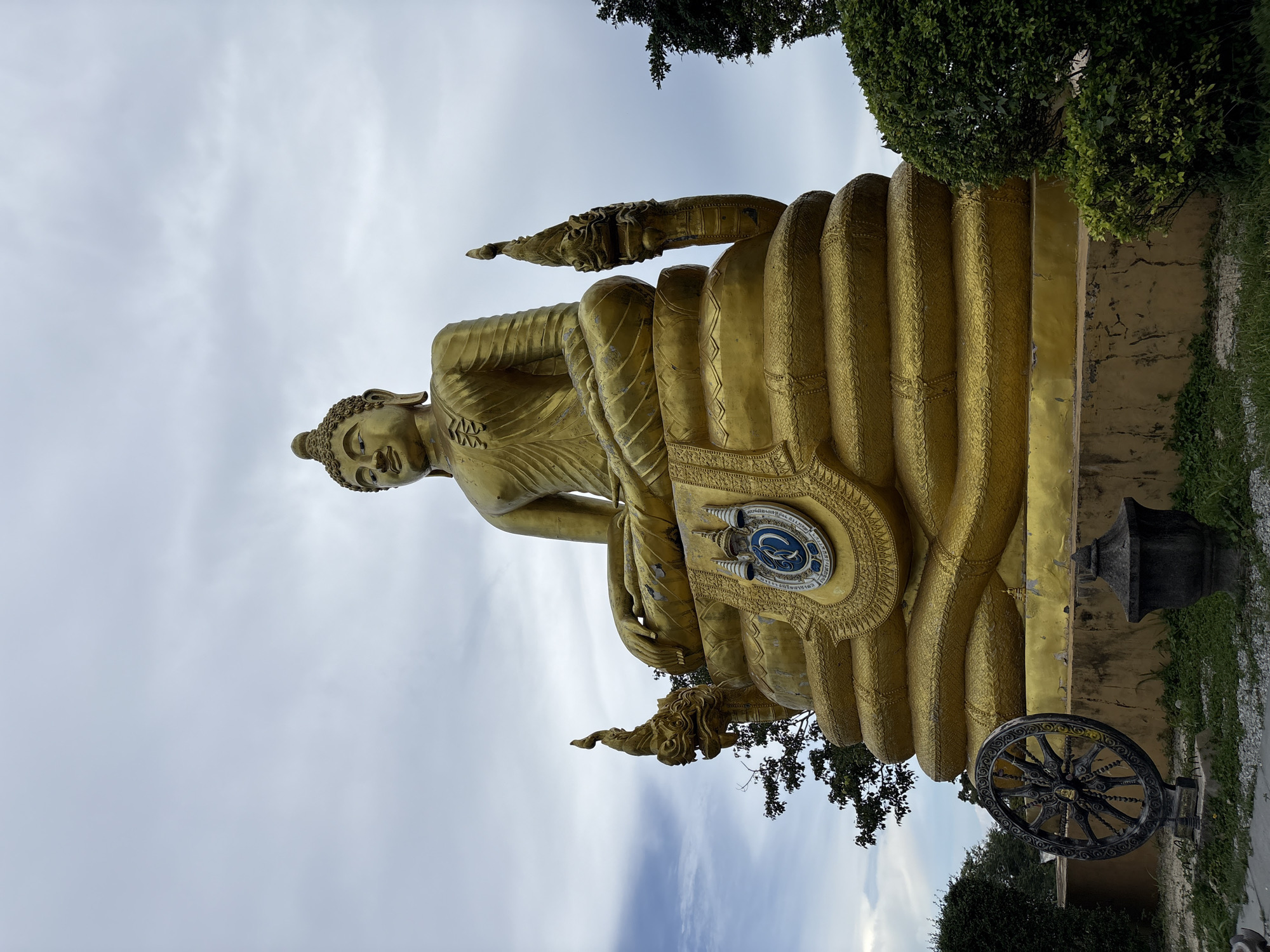 A large, golden Buddha statue sits outdoors in a seated position, with a cloudy sky overhead. The statue is adorned with intricate details, including what appear to be Naga heads on either side of its base. A circular emblem with blue and white colors is visible on the front of the base. In the foreground, to the left of the statue, is a dark, ornate wheel-like object, possibly a dharma wheel, and a small dark urn or vessel. Lush green bushes and trees surround the base of the statue. The overall scene suggests a temple or sacred site in Rawai, Thailand, given the presence of the Buddha statue and decorative elements. The lighting suggests it is daytime, with diffused light due to the cloud cover.