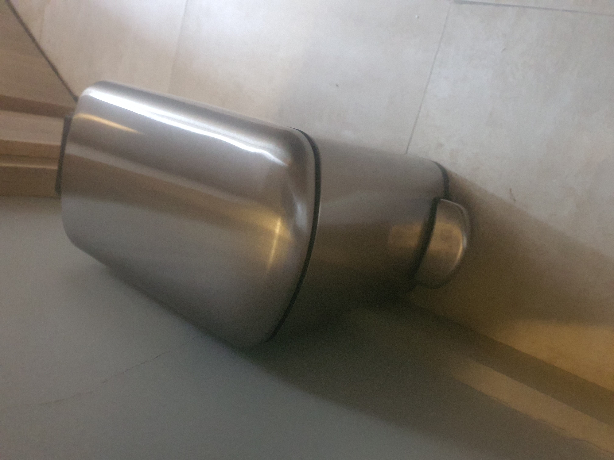 The image shows a close-up, angled view of a metallic, cylindrical object lying on a tiled floor. The object is silver and has a smooth, reflective surface. It appears to be a single piece with a rounded end. The background consists of light-colored tiles, with some visible grout lines. To the left of the object, there are some wooden planks stacked horizontally, suggesting a construction or storage area. The lighting in the image is bright, casting reflections on the metallic object and illuminating the scene. There is no visible text in the image. The overall impression is one of a simple, static composition focusing on the texture and form of the metallic object within a neutral setting.