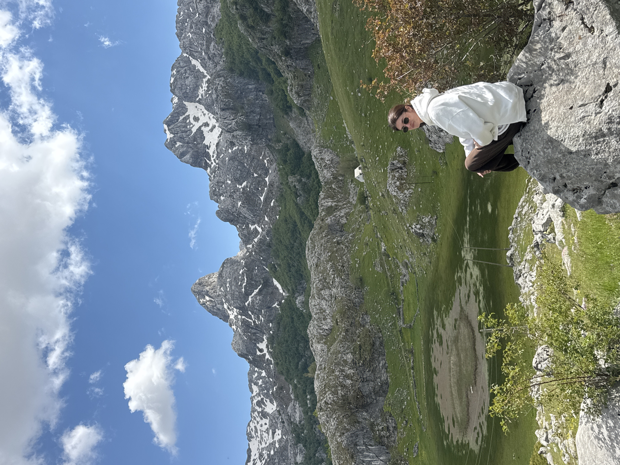 A slightly rotated, eye-level outdoor shot in Brskut, Montenegro, shows a person sitting on a rocky outcrop overlooking a valley with snow-capped mountains in the background, under a blue sky with scattered clouds.
The person is wearing sunglasses, a white hoodie, and dark pants, and is crouched on a light grey rock formation on the right side of the frame. They are looking toward the valley, which is lush with green grass and contains an oval shaped, dried up or shallow lake or depression. A few small, indistinct buildings are visible in the valley. Power lines extend vertically across the valley.
On the left side of the frame is a mountain range with grey, rocky peaks speckled with snow. The sky above is a bright blue with scattered white clouds. The perspective is that of a wide shot, showing a panoramic view of the mountainous terrain.
