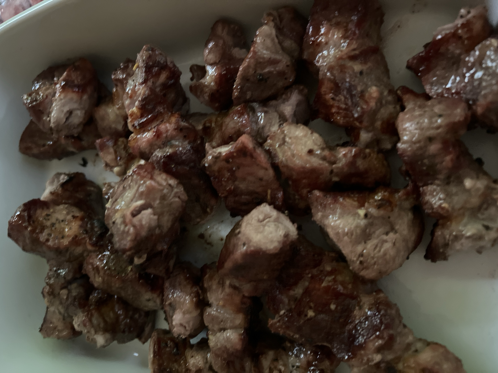 A close-up, overhead shot showcases a white rectangular dish filled with bite-sized pieces of cooked meat. The meat appears to be seared, with some pieces exhibiting a darker brown crust and others a lighter pinkish-brown interior. Some pieces have visible flecks of black pepper. The dish is set against a plain, light-colored background, suggesting it might be a kitchen counter or dining table. No individuals or specific locations within Revelstoke, Canada are visible in the image.