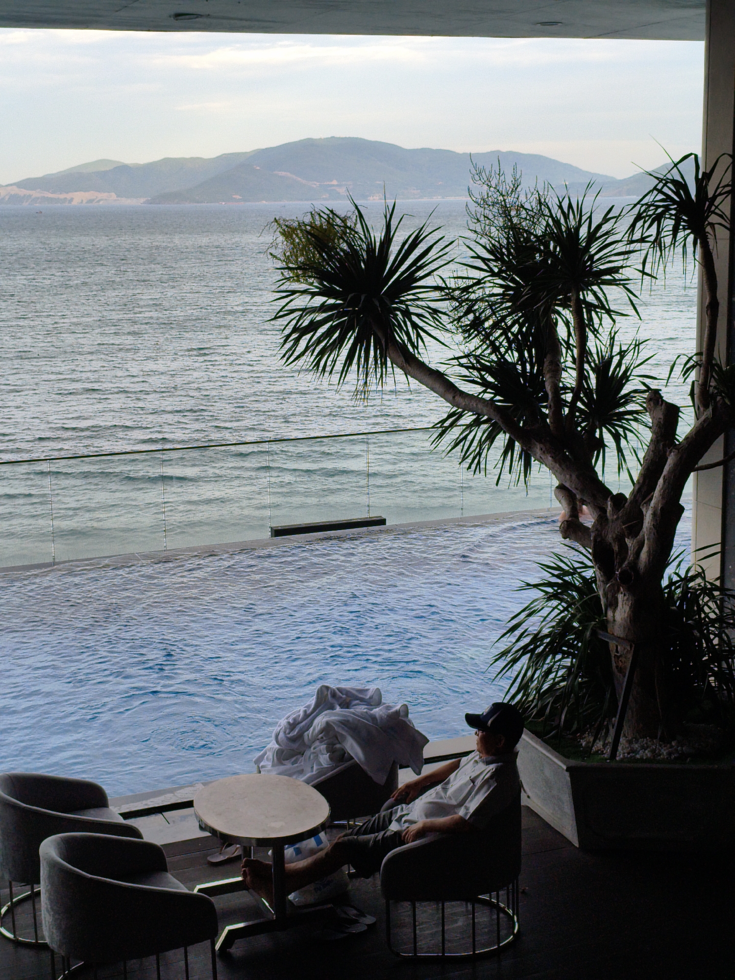 An outdoor scene in Nha Trang, Vietnam, shows a man in a light-colored short-sleeved shirt and dark pants, wearing a baseball cap and sunglasses, relaxing in a chair next to a small round table. He is seated by a swimming pool with a glass railing overlooking the sea. In the foreground, two empty chairs and the table with a folded white towel on it are visible. A tree with spiky leaves shades part of the scene. In the background, a tranquil sea stretches to the horizon, with hazy, layered mountains in the distance under a pale sky. The lighting suggests it is late afternoon or early morning.